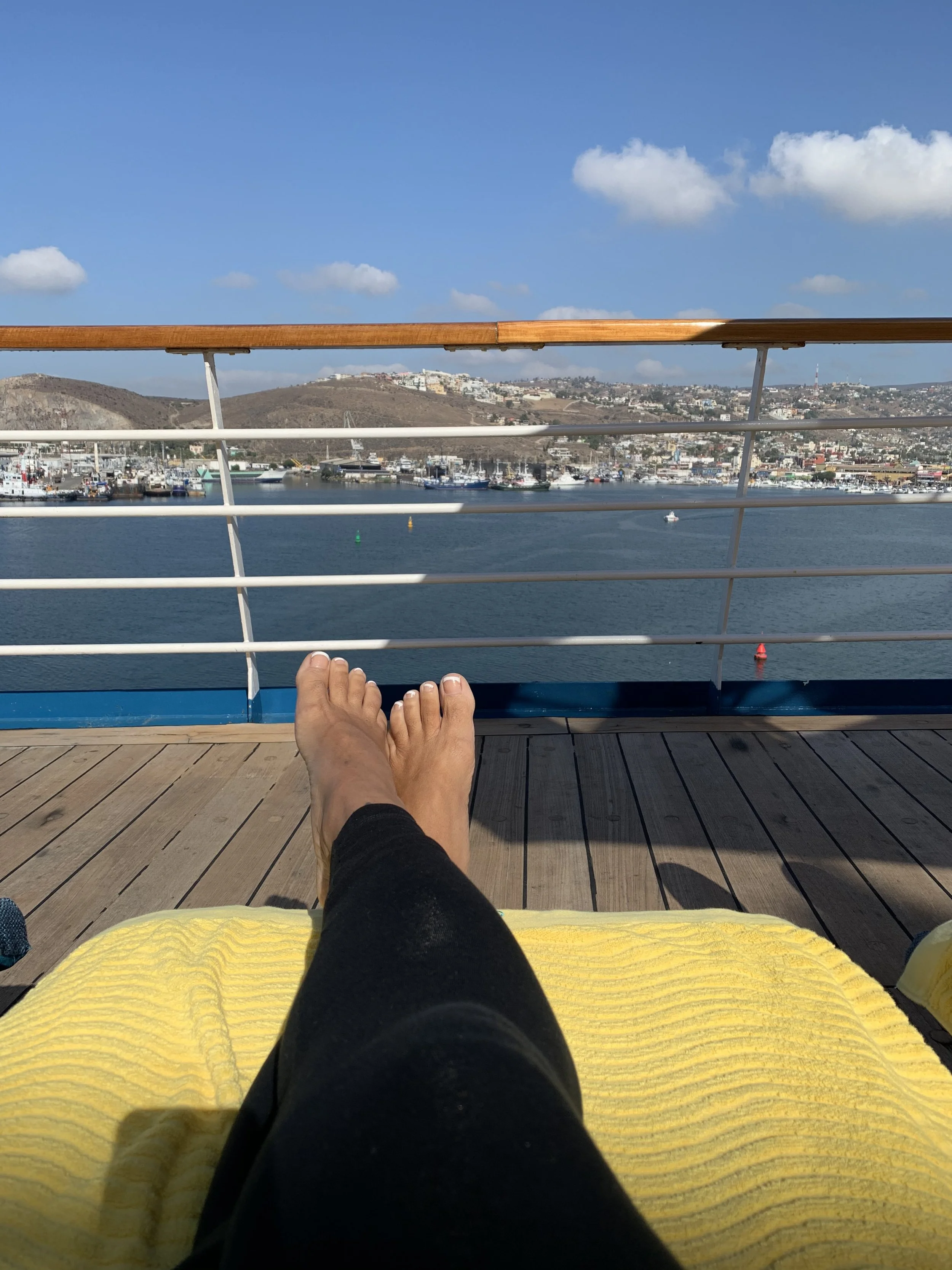 Person relaxing on a boat deck with legs stretched out, overlooking a marina with boats and hills in the background, under a partly cloudy sky.