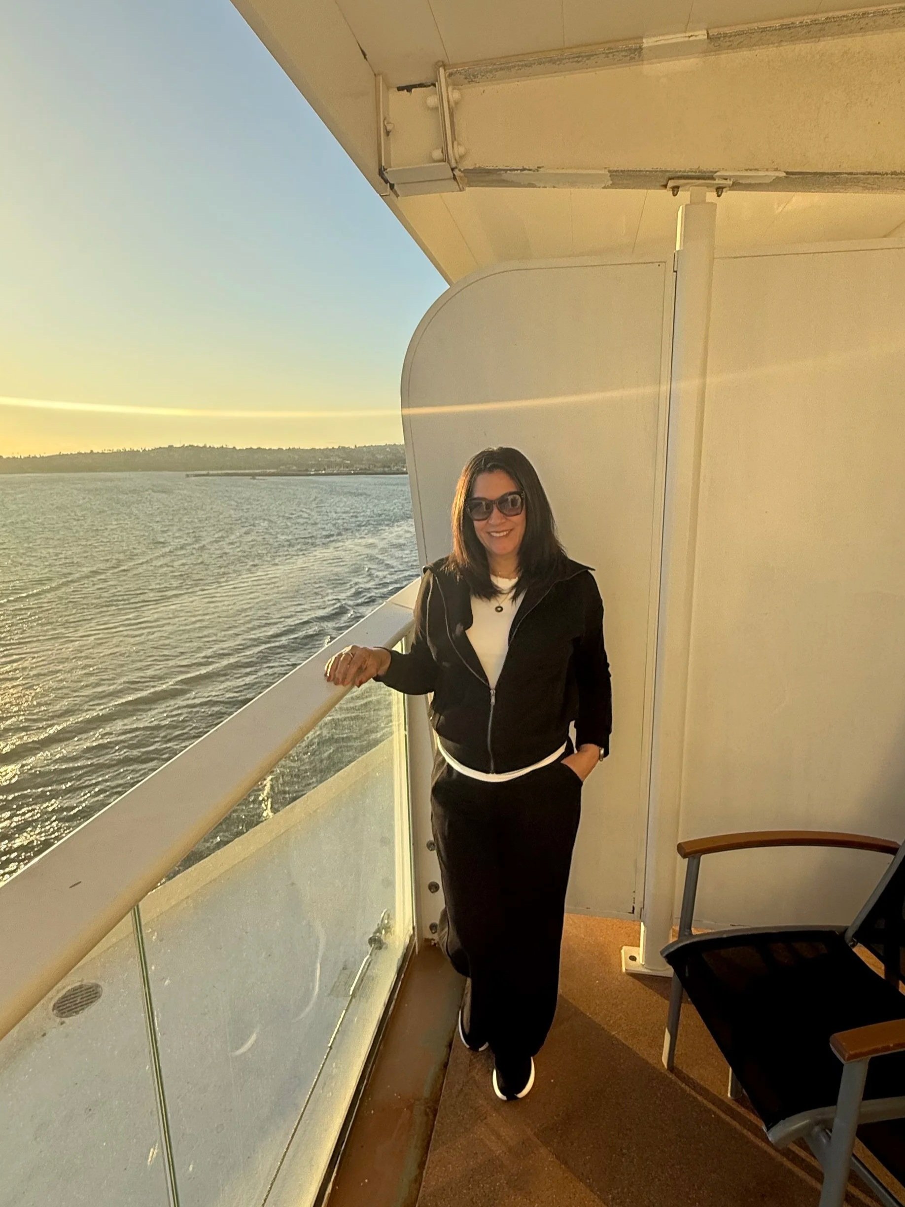 A woman in black clothing and sunglasses on a boat, standing on a balcony with water and a distant shoreline in the background during sunset.