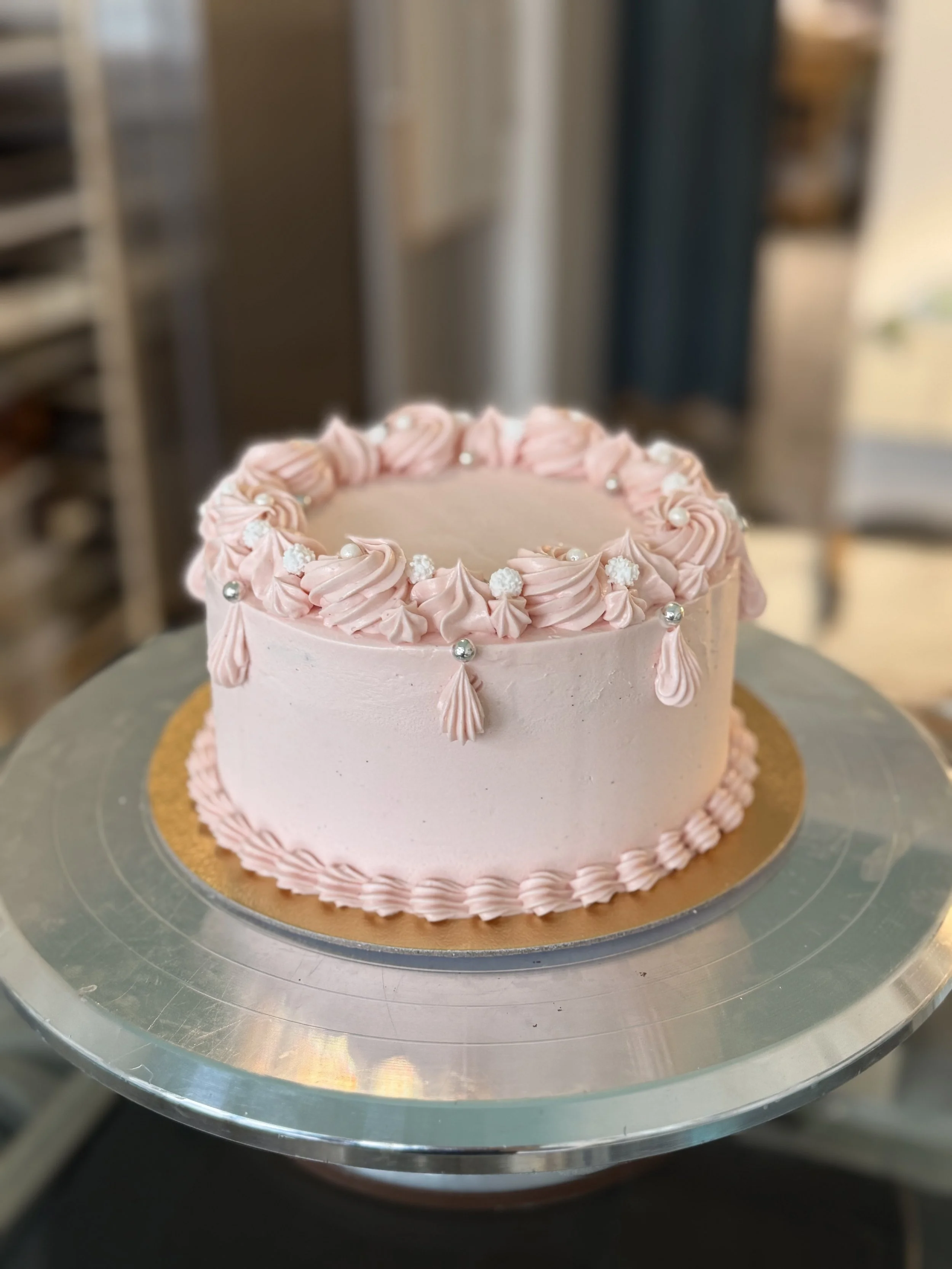 Pink frosted birthday cake decorated with piped frosting swirls, white sugar pearls, and edible decorations, sitting on a cake stand in a kitchen.