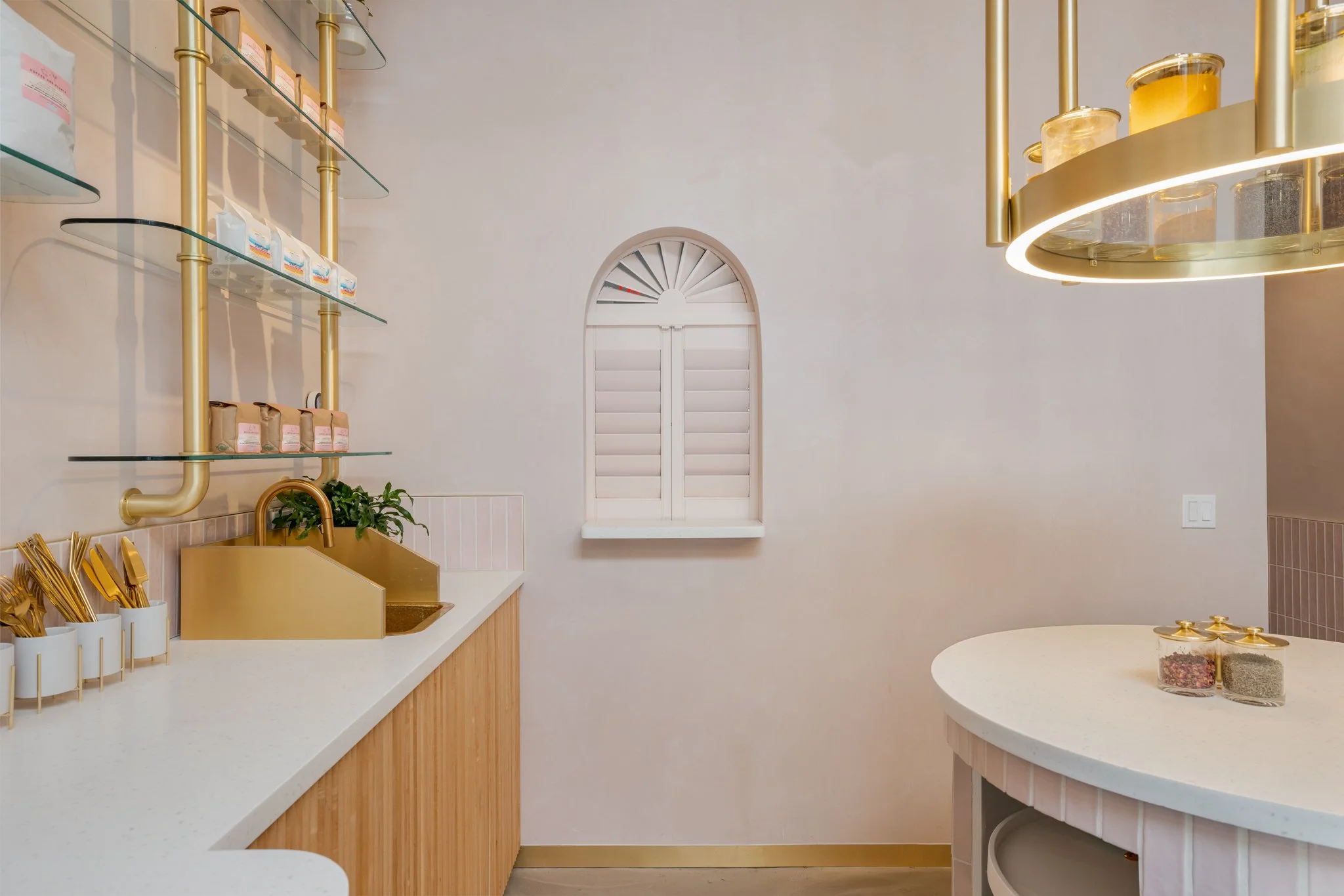 Minimalist kitchen with white walls and countertops, gold accents on the sink, shelves, and chandelier, and a small window with white shutters.