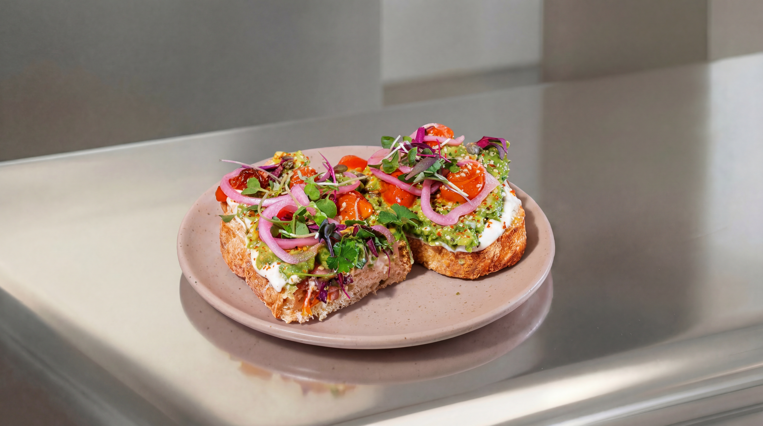 Open-faced sandwich on a pink plate with avocado, cherry tomatoes, pickled onions, greens, and sauce on toasted bread.