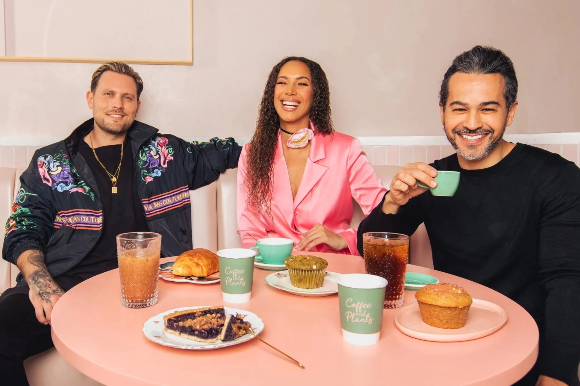 Three people sitting at a pink table in a café, smiling and enjoying coffee with pastries and pie.