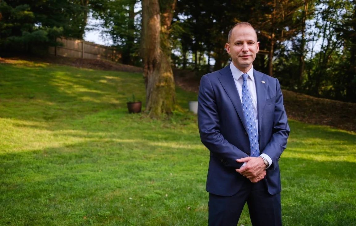 Photo of candidate Jonathan Estreich, standing in a grassy area with his arms folded and looking at the camera. He's wearing a blue suit and light blue tie. He has a US Flag pin on his lapel.
