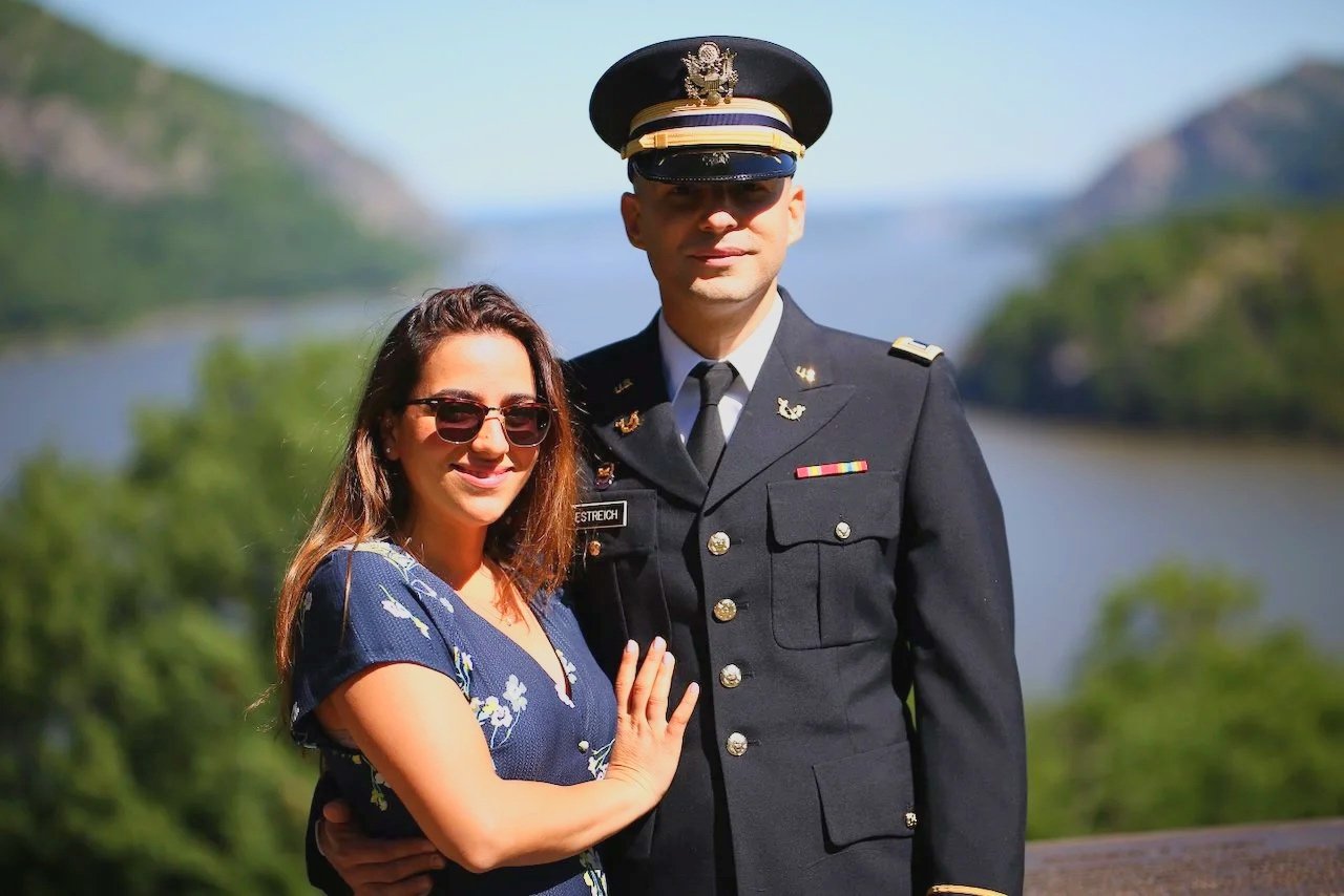 Jon Estreich and his wife standing on a bluff at West Point following his promotion ceremony to the rank of Major in the US Army. There is a river and mountain in the background.