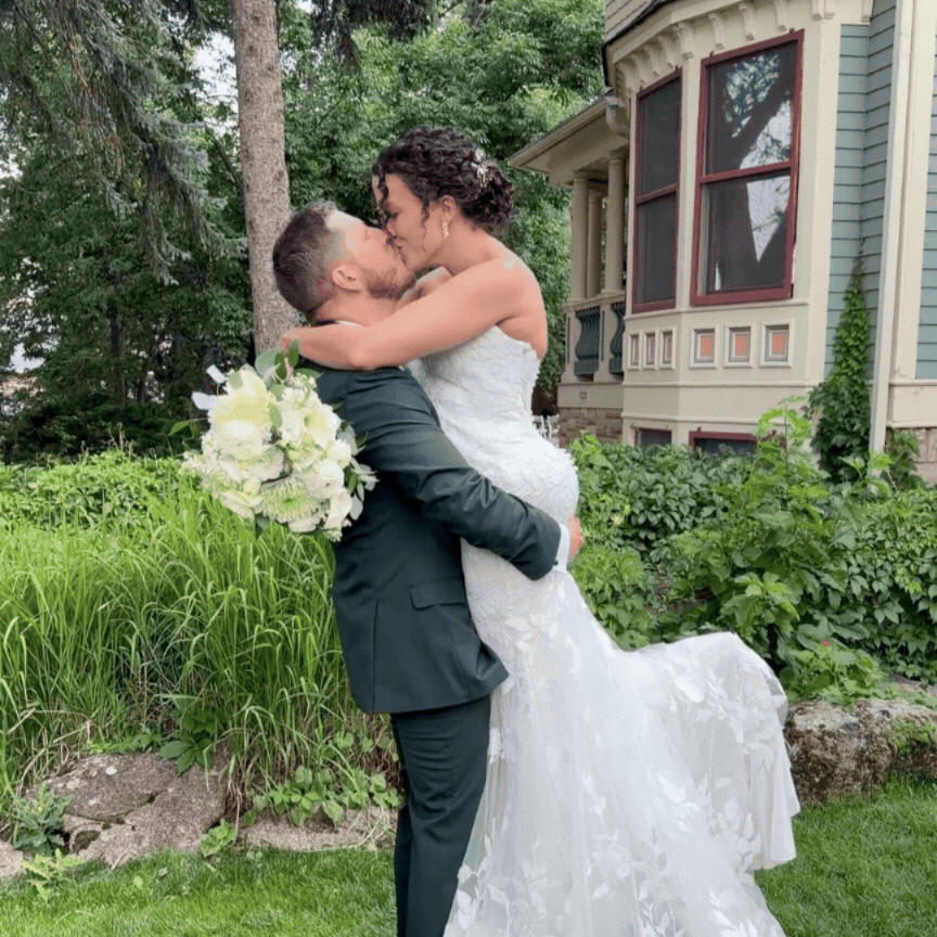 A newlywed couple kissing outdoors, with the groom lifting the bride, holding a bouquet of white flowers, against a garden and house background.