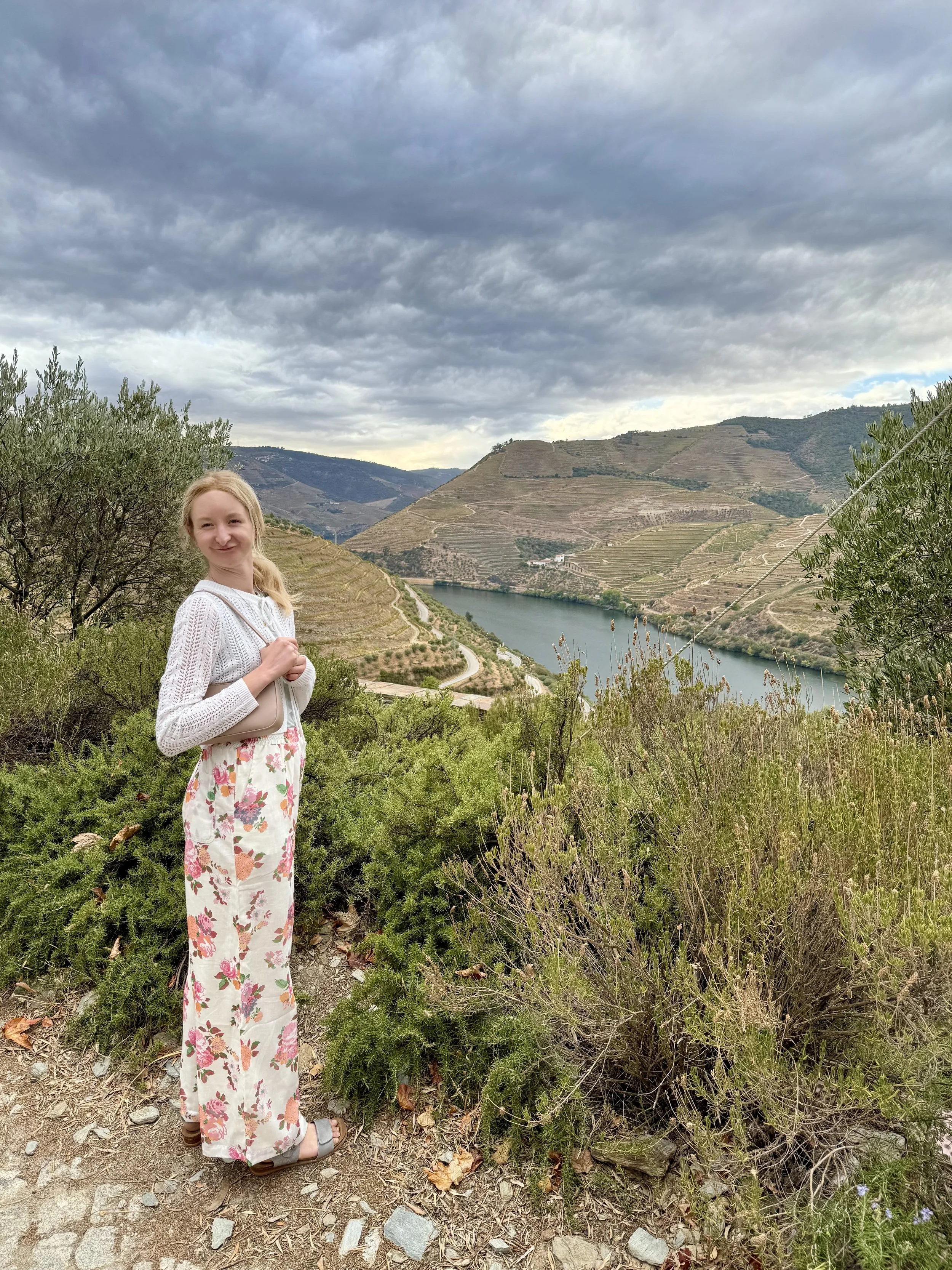 A young woman standing on a hillside with a river and terraced hills in the background under a cloudy sky.