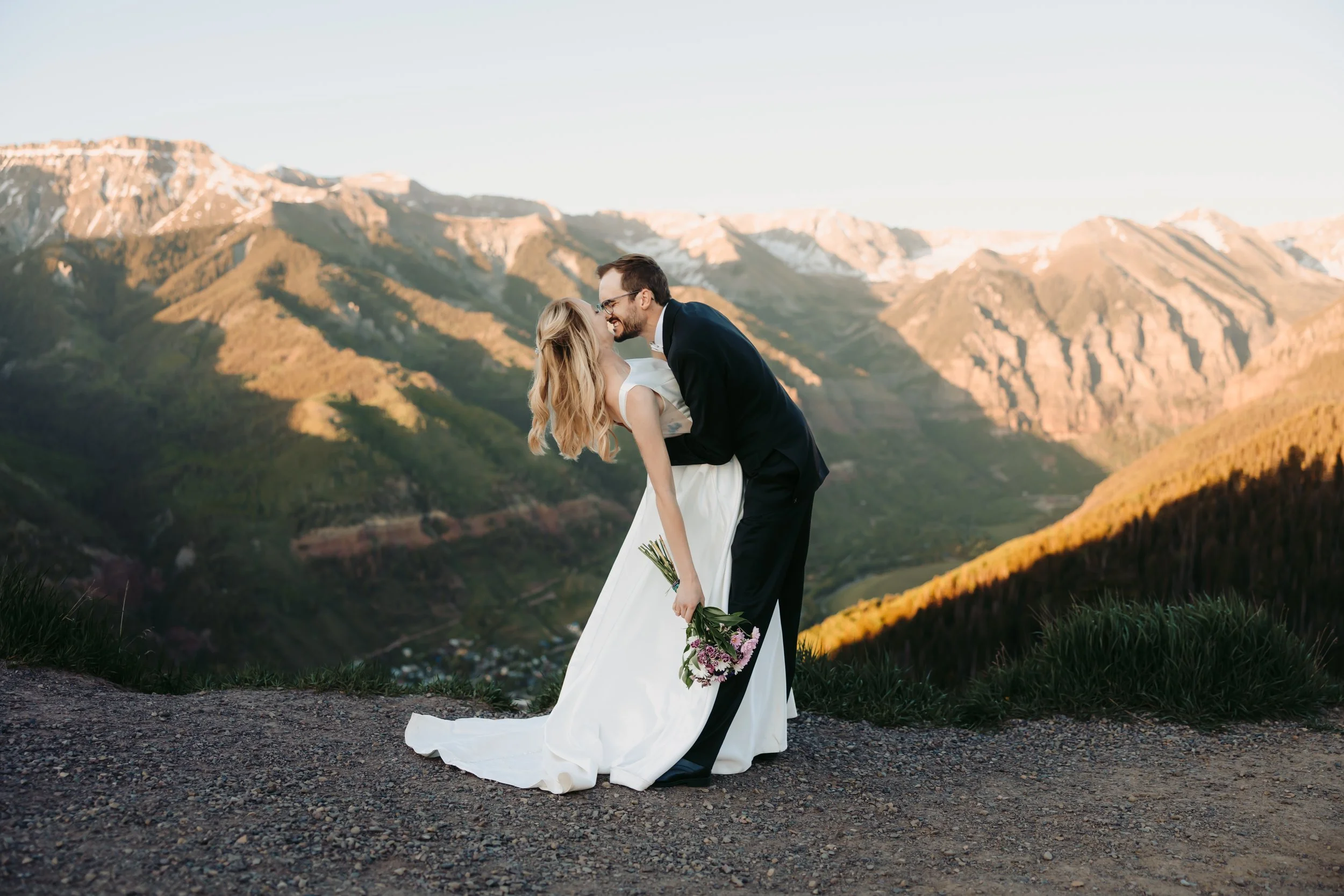 A newlywed couple sharing a romantic dance outdoors at sunset against a mountain landscape. The bride is in a white wedding dress holding a bouquet, and the groom is in a black suit. They are smiling and leaning close to each other.