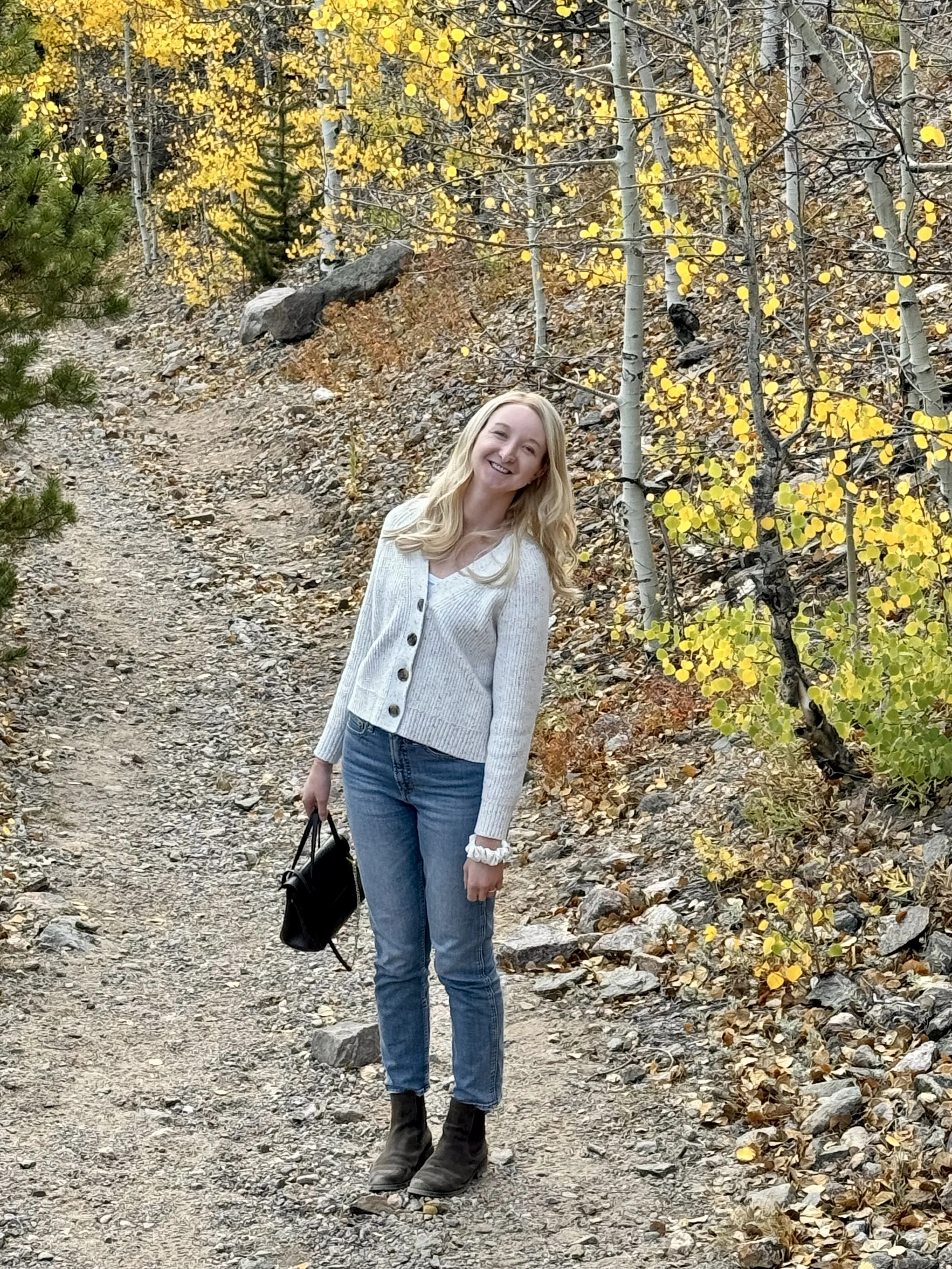 A woman with long blonde hair wearing a white sweater, blue jeans, and black boots, standing on a dirt trail in a forest with yellow autumn leaves on the trees and ground.