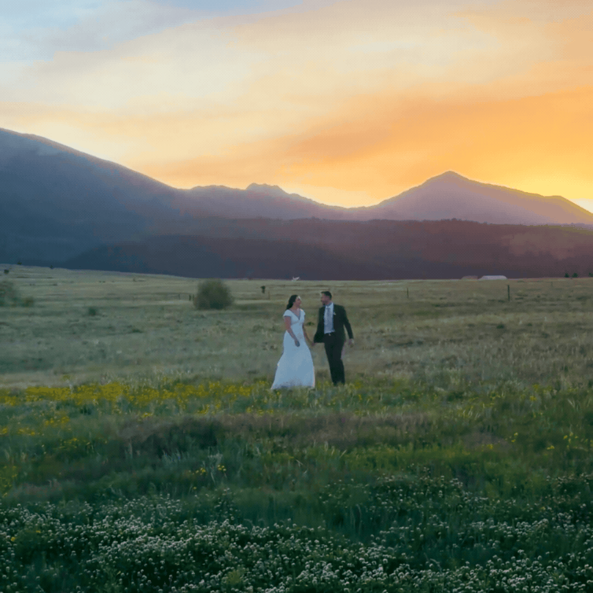 A bride and groom holding hands in a grassy field at sunset with mountains in the background.