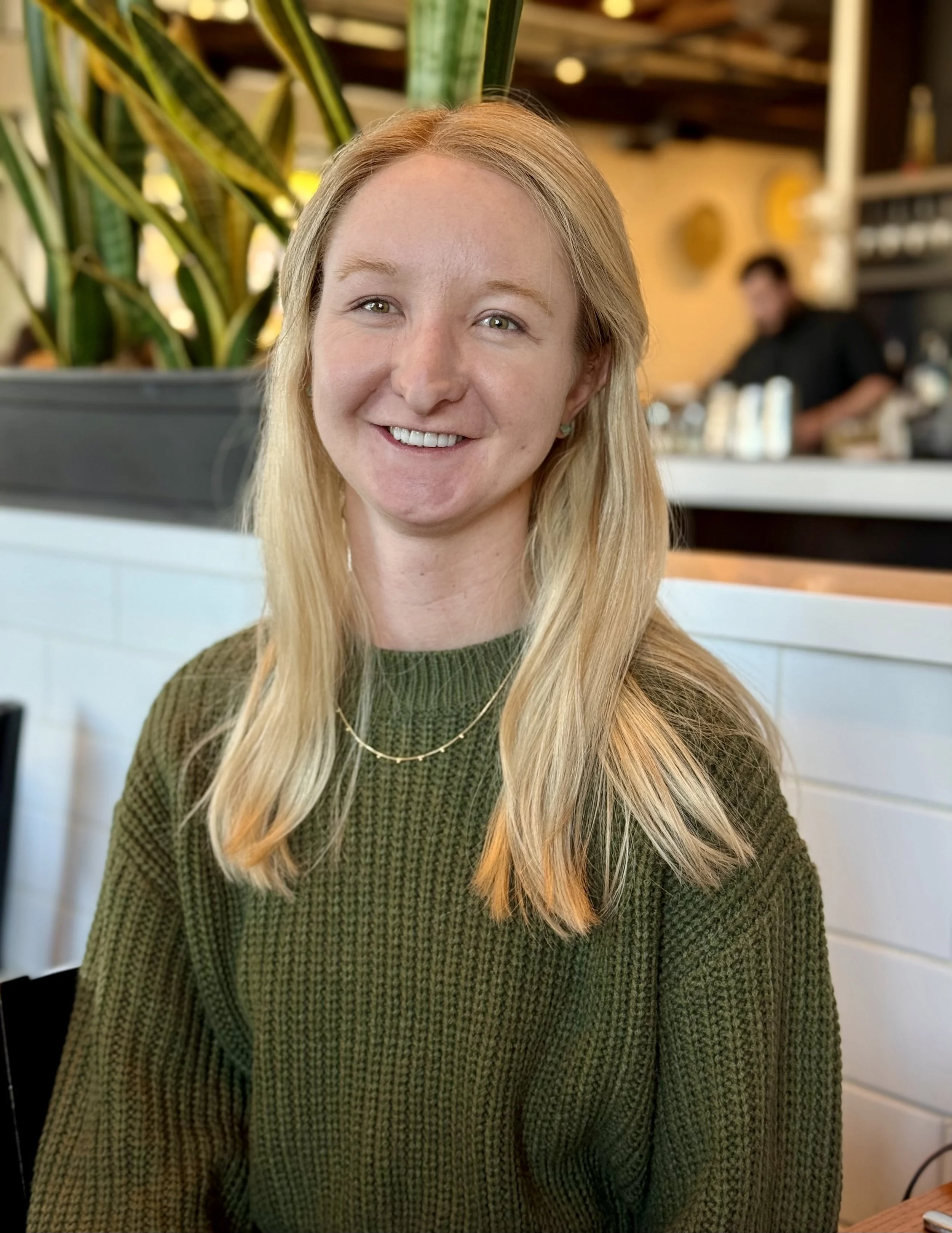 A smiling young woman with long blonde hair wearing a green sweater and a gold necklace, sitting in a restaurant or cafe.