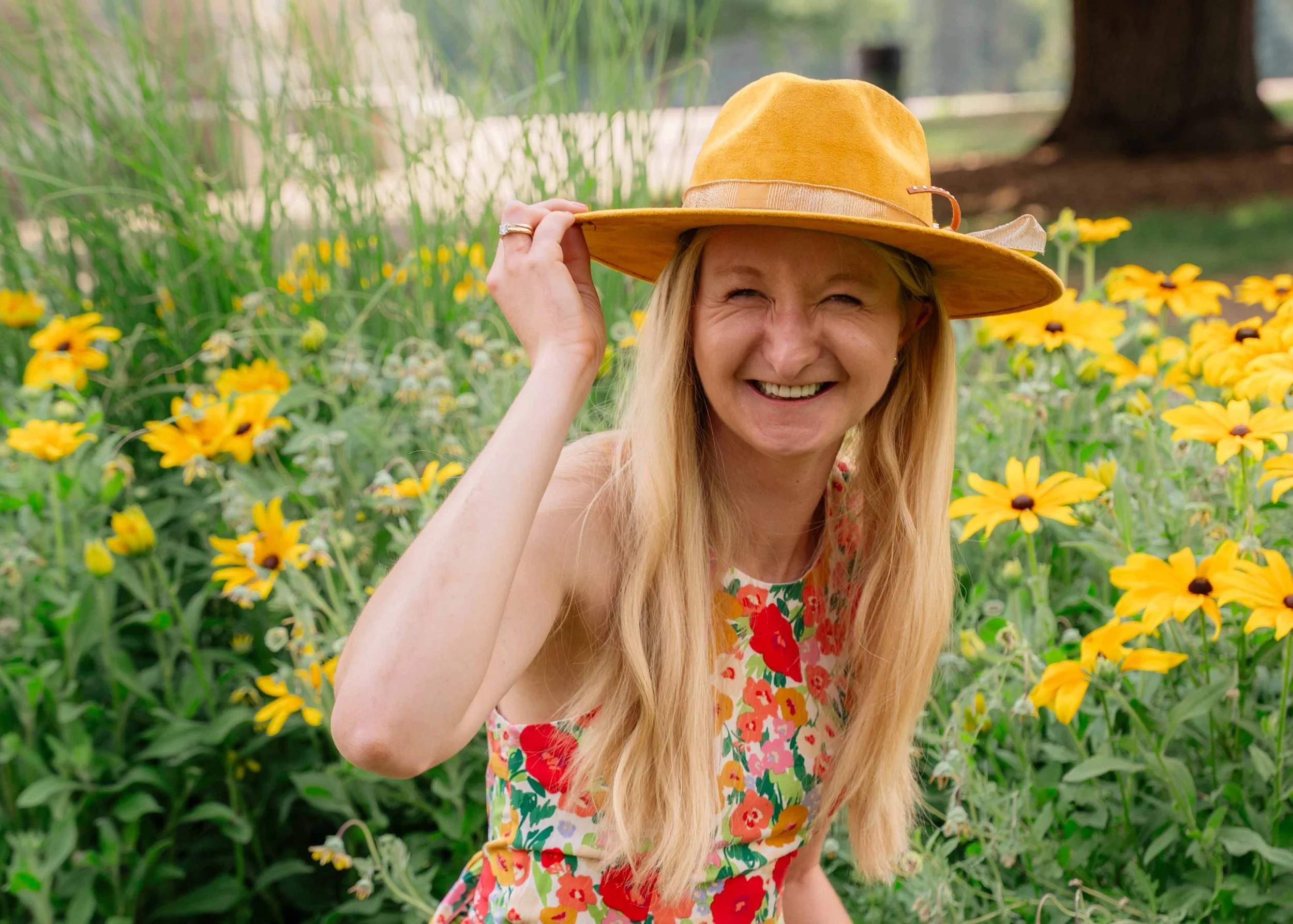 A woman with long blonde hair smiling and winking while wearing a wide-brimmed yellow hat, standing among yellow flowers and green plants in a park or garden setting.