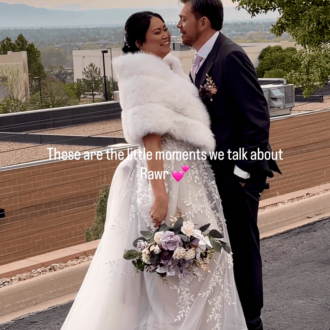A bride and groom on a rooftop, smiling and looking at each other, with the bride holding a bouquet of flowers. They are dressed in wedding attire, with the bride wearing a white dress and fur shawl, and the groom in a suit. There are city buildings and trees in the background.