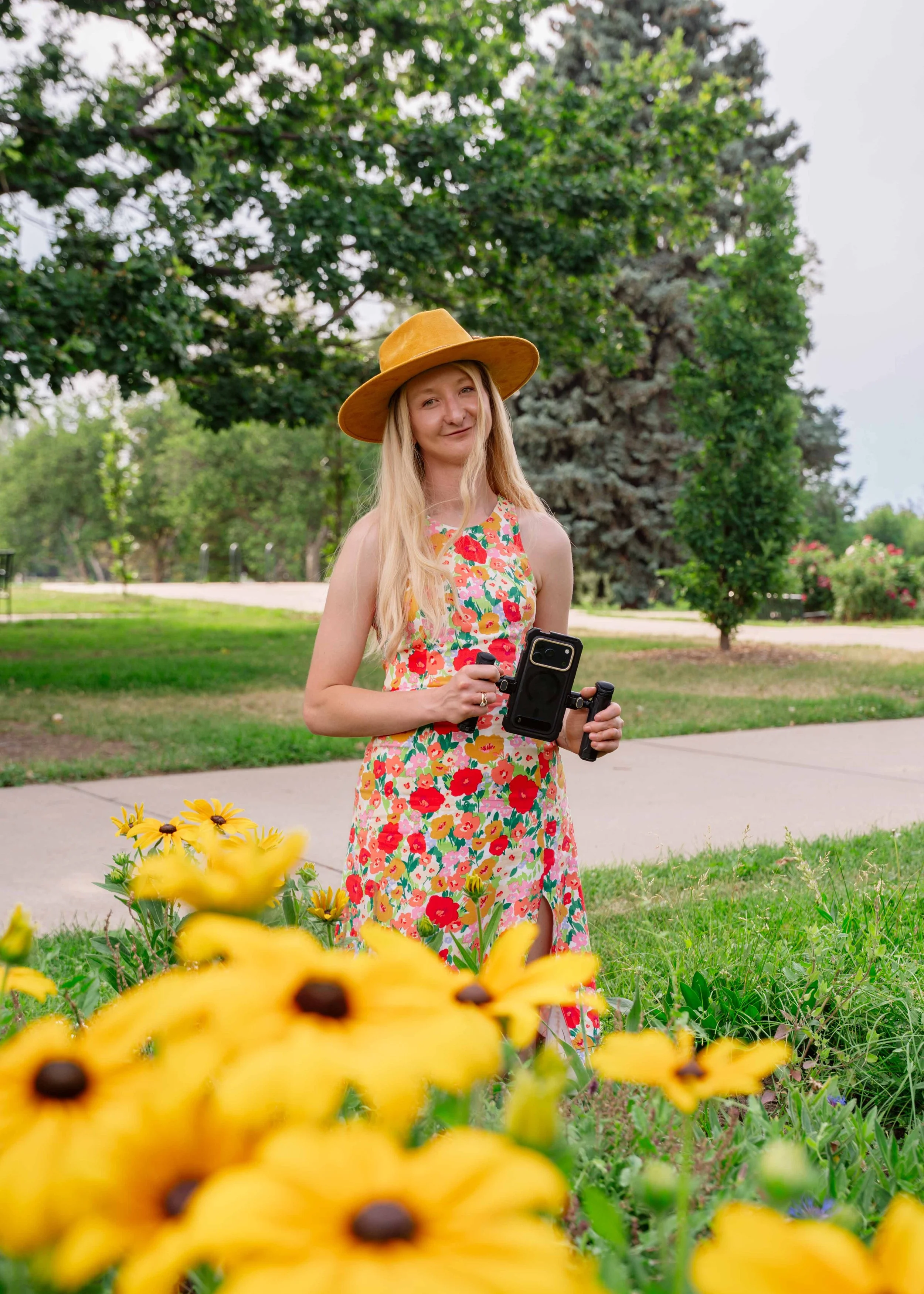 Woman in a floral dress and wide-brimmed hat holding a phone, standing near yellow flowers in a park with trees in the background.