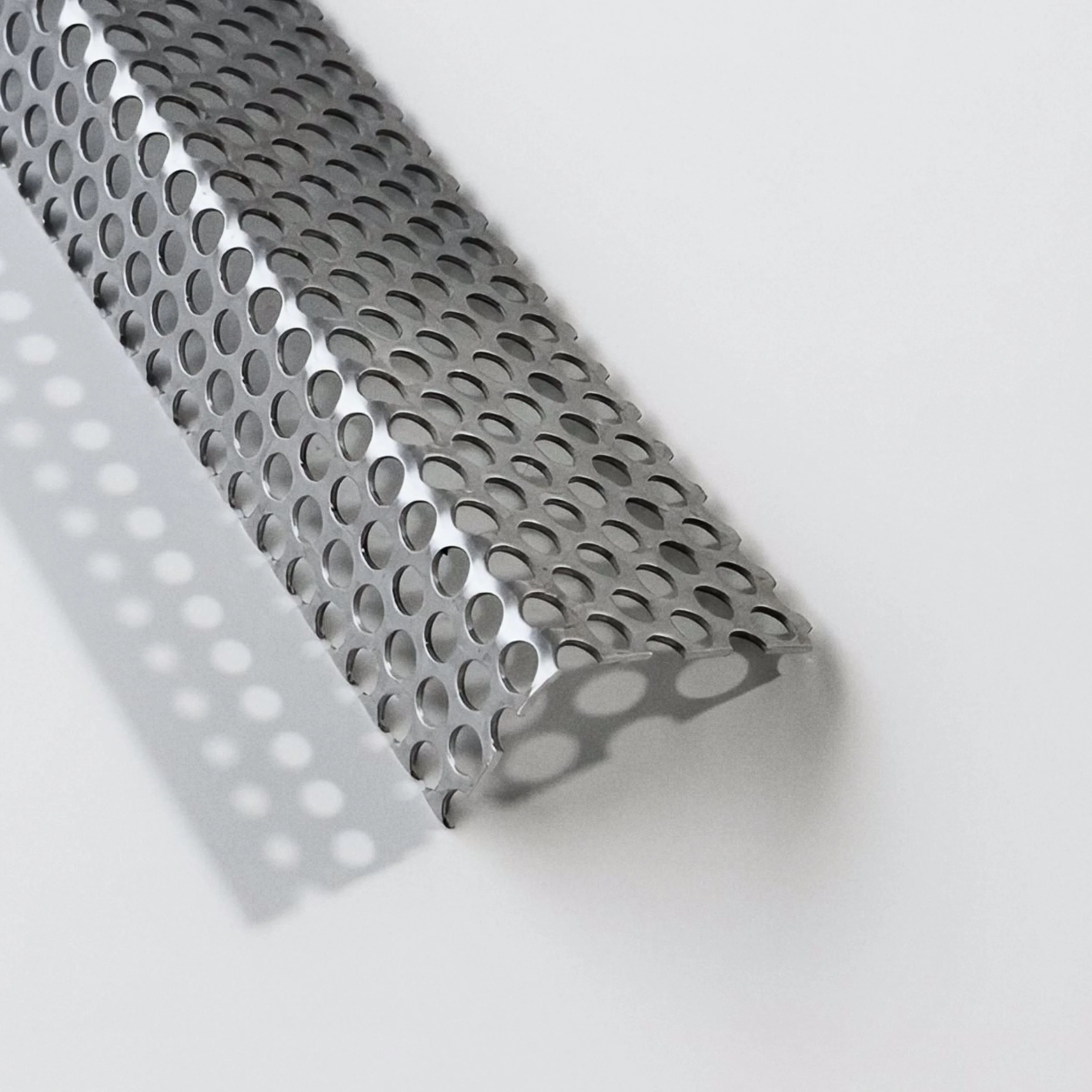 A close-up of a metallic, perforated sheet with evenly spaced oval holes, placed on a white surface with soft shadowing.
