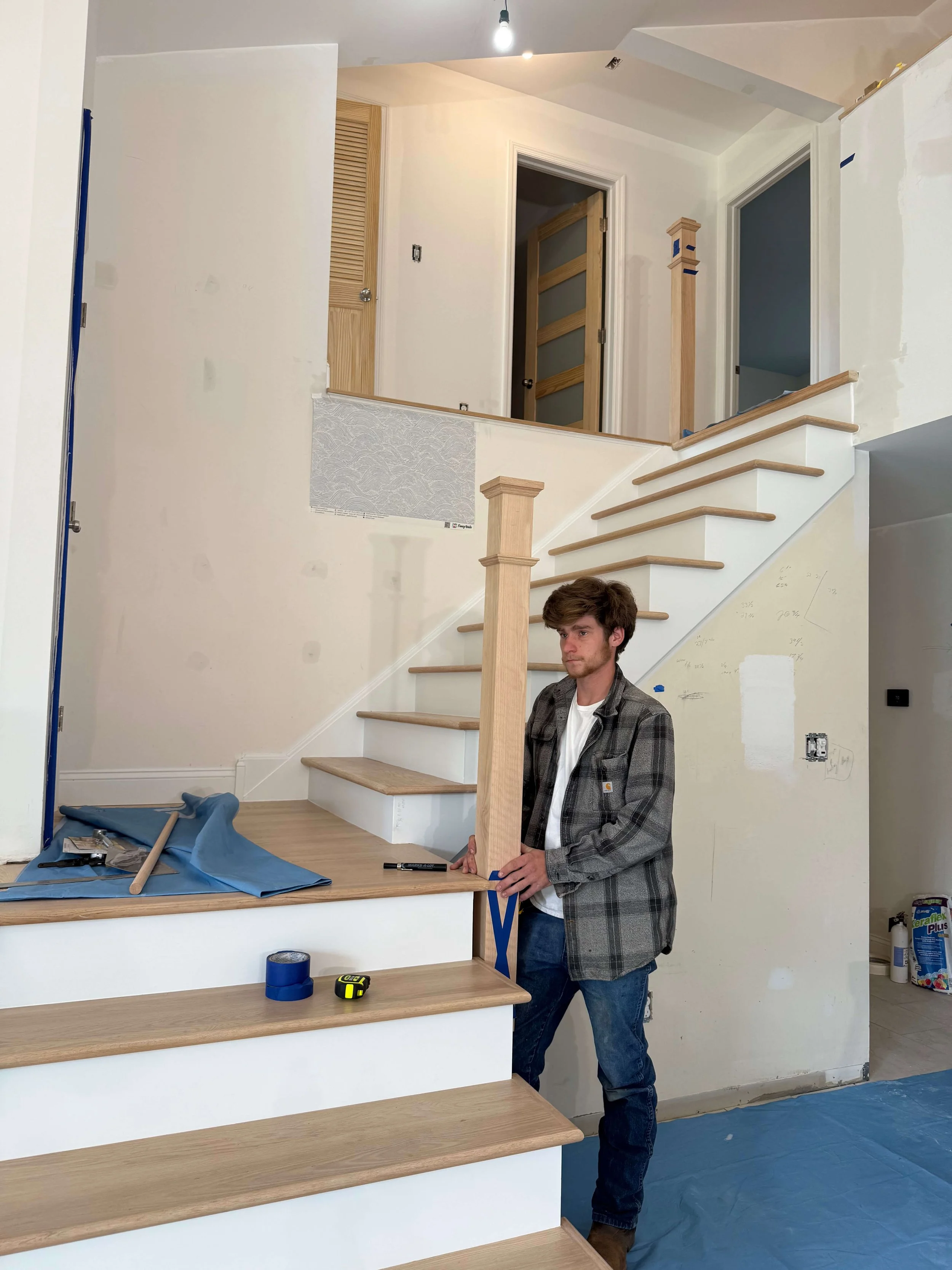A young man in a plaid jacket holding a measuring tape stands next to wooden stairs during a home renovation. The stairs are partially built, and work tools are on the counter. The interior walls are in the process of being painted or finished.