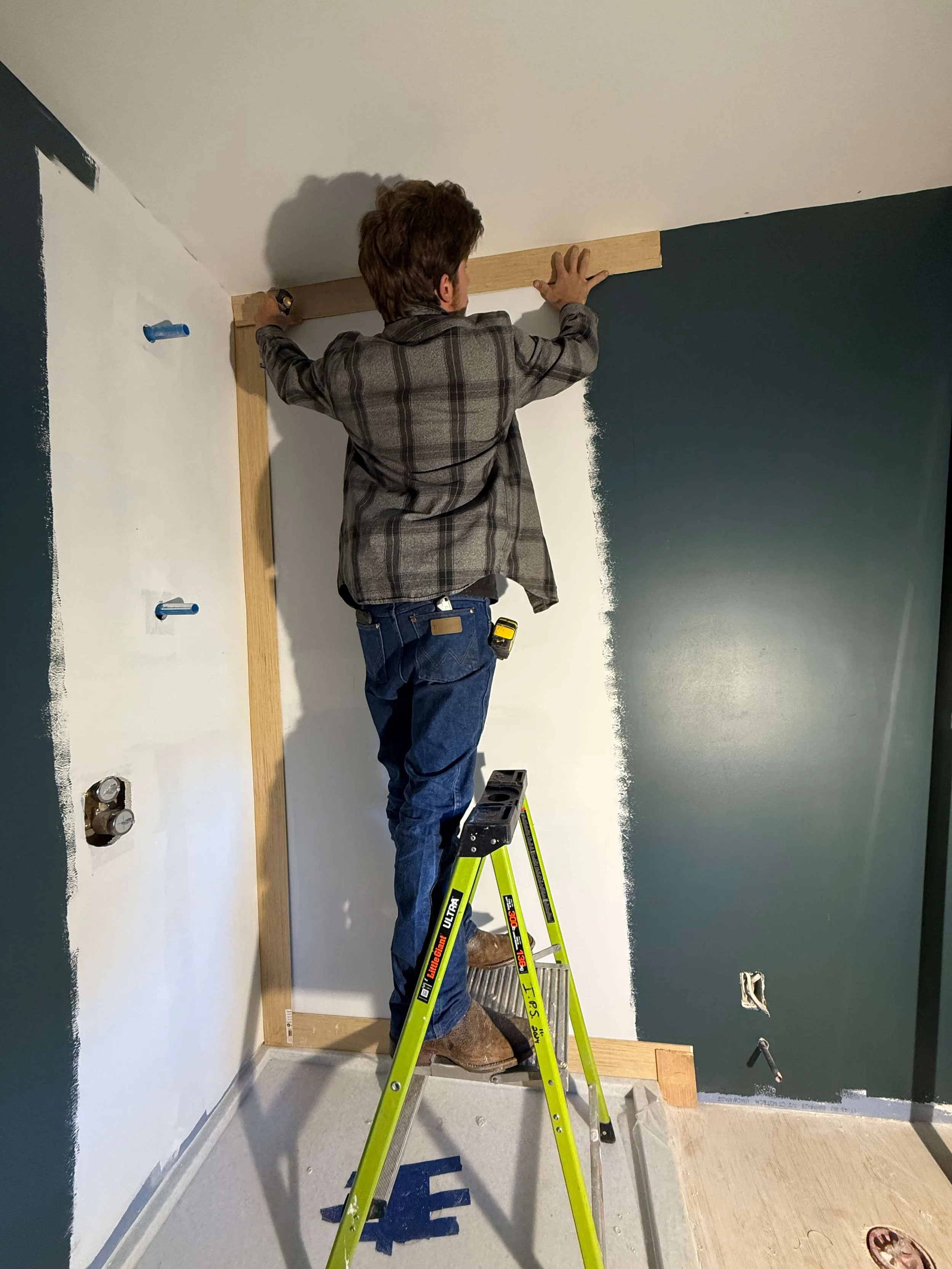 A man is standing on a step ladder painting the top part of a wall, which is dark green, in a room with a white ceiling and light-colored flooring.