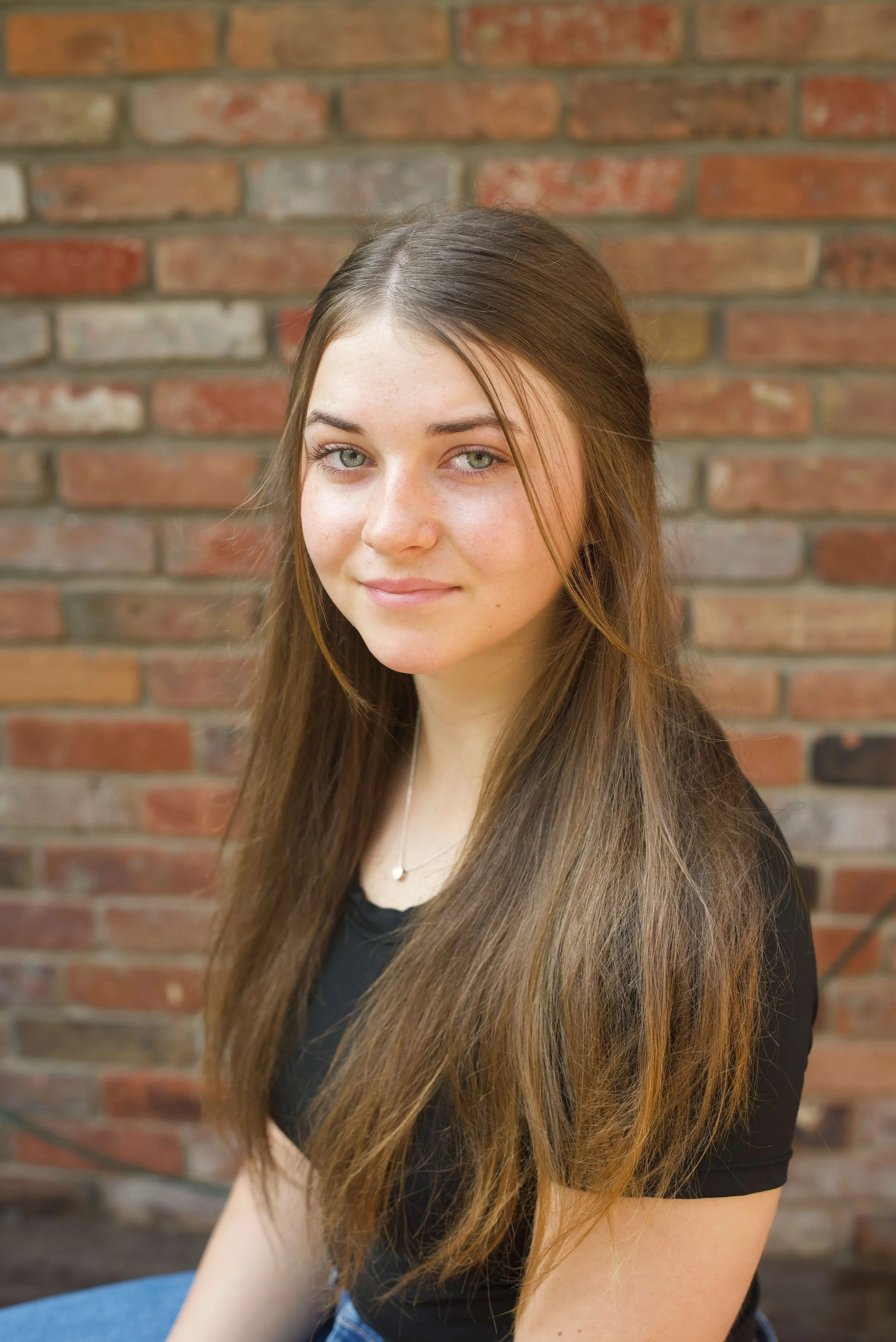 Young woman with long brown hair wearing a black t-shirt in front of a red brick wall.