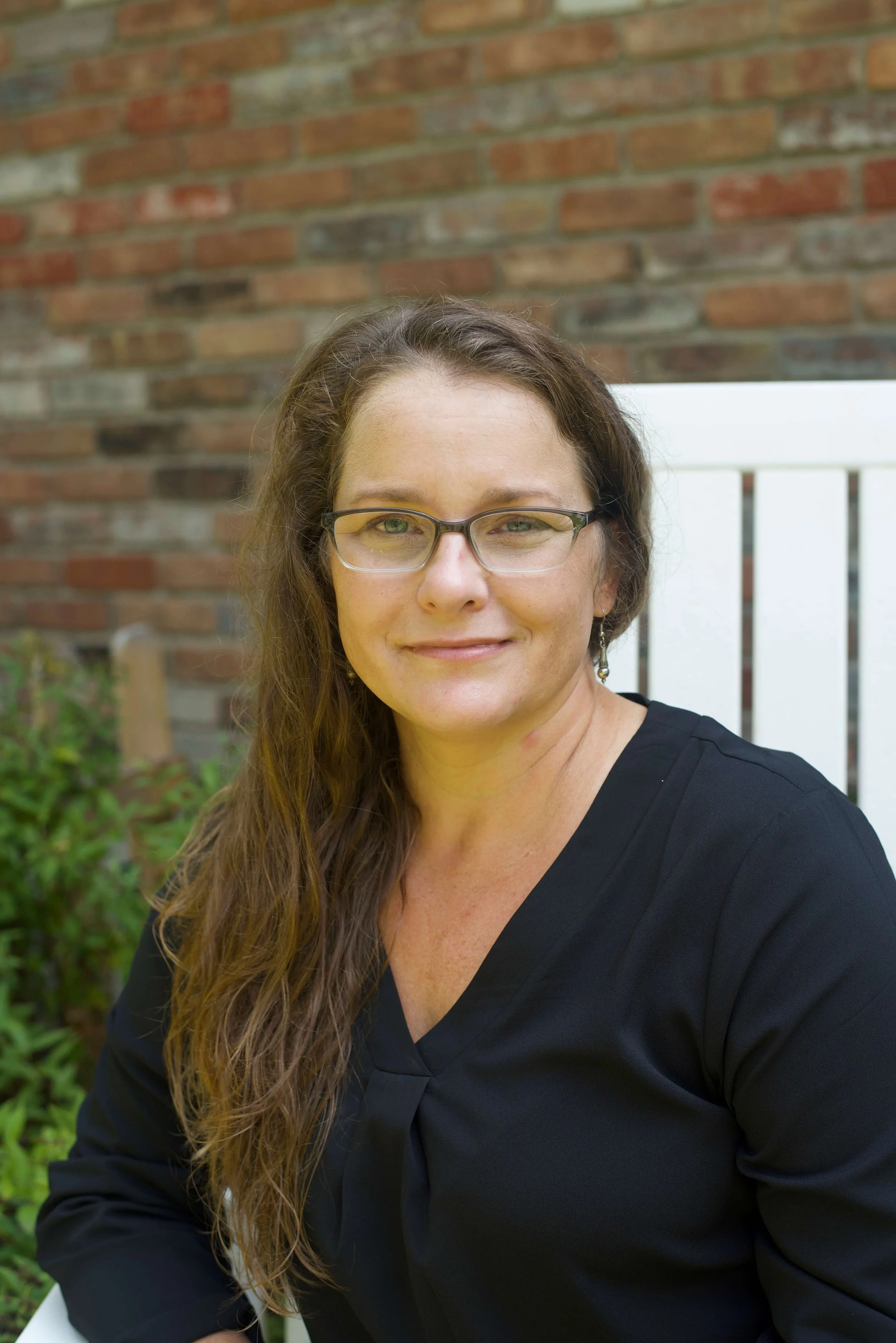 A woman with long brown hair and glasses, wearing a black top, smiling outdoors in front of a brick wall and greenery.