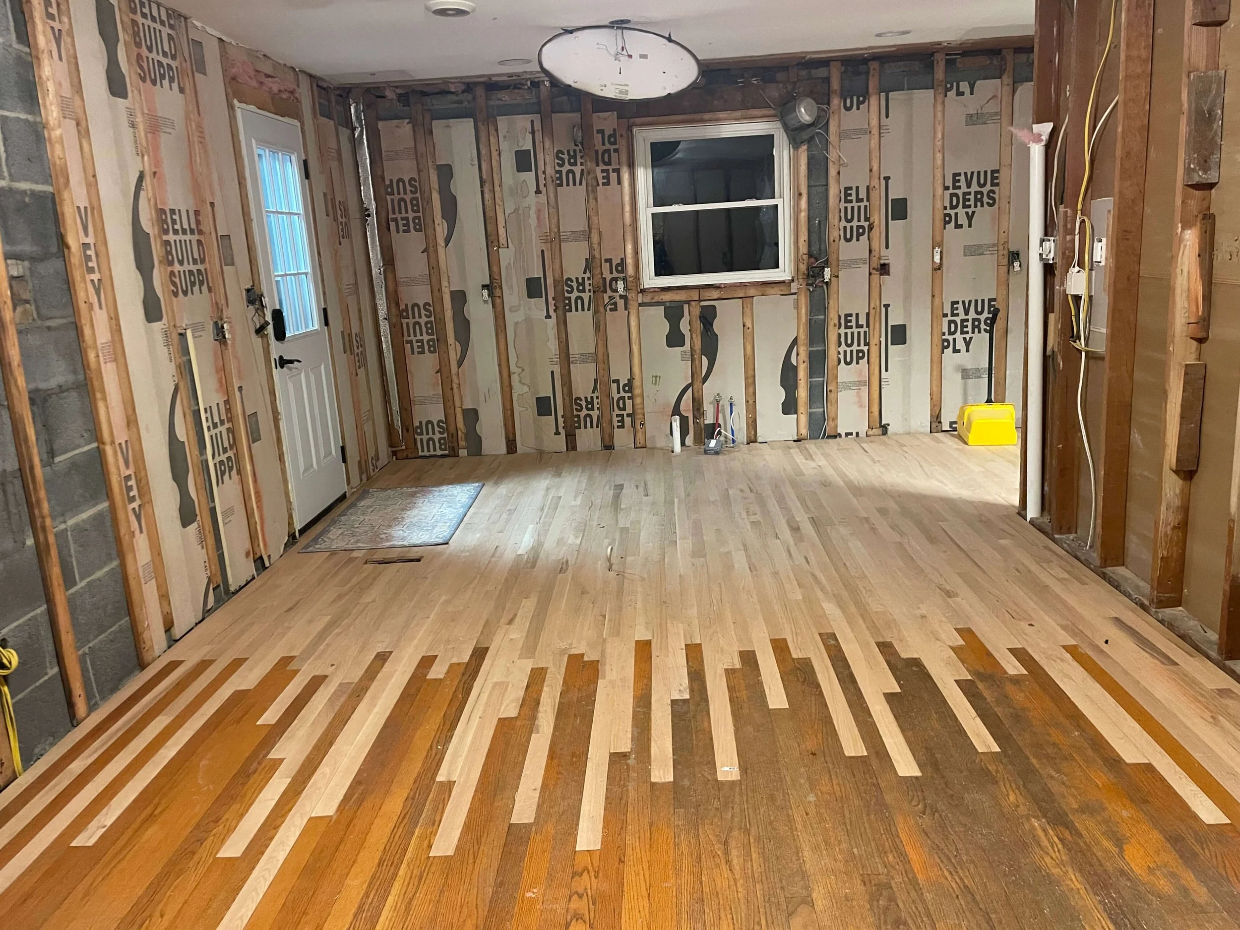 Room under renovation with exposed wooden wall studs, window, white door, and a new wooden floor with patterned design. Electrical wiring and outlets are visible.