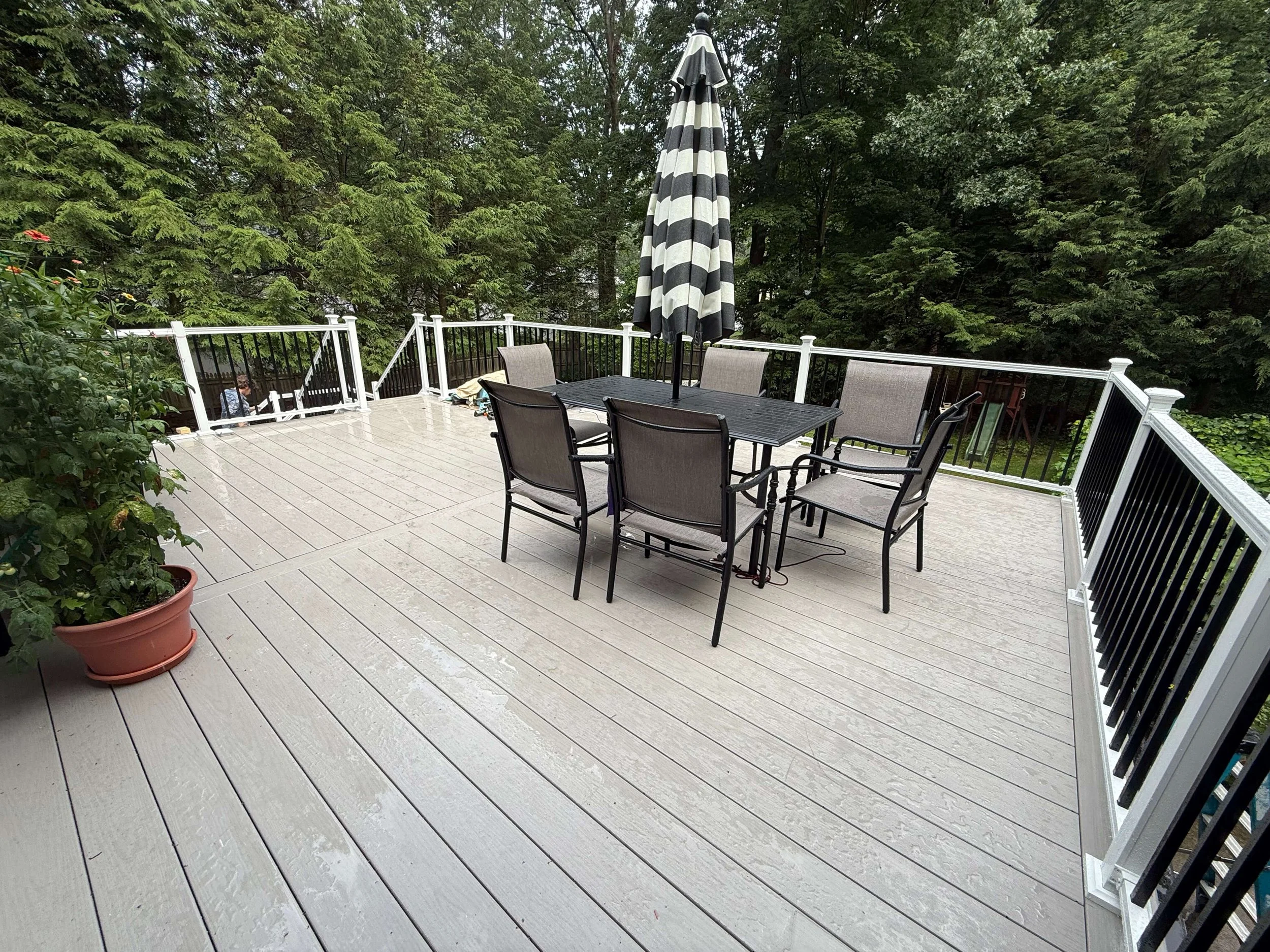 A wooden outdoor deck with a patio table and chairs, a black and white striped umbrella, some potted plants, and a surrounding green forest.