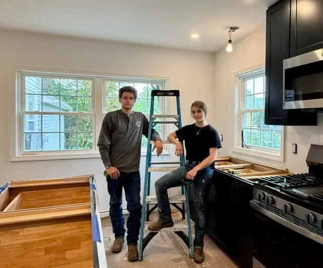 Two people in a kitchen with renovation in progress, one person standing next to a ladder and the other sitting on the ladder's step. There are windows, a stove, and construction materials present.