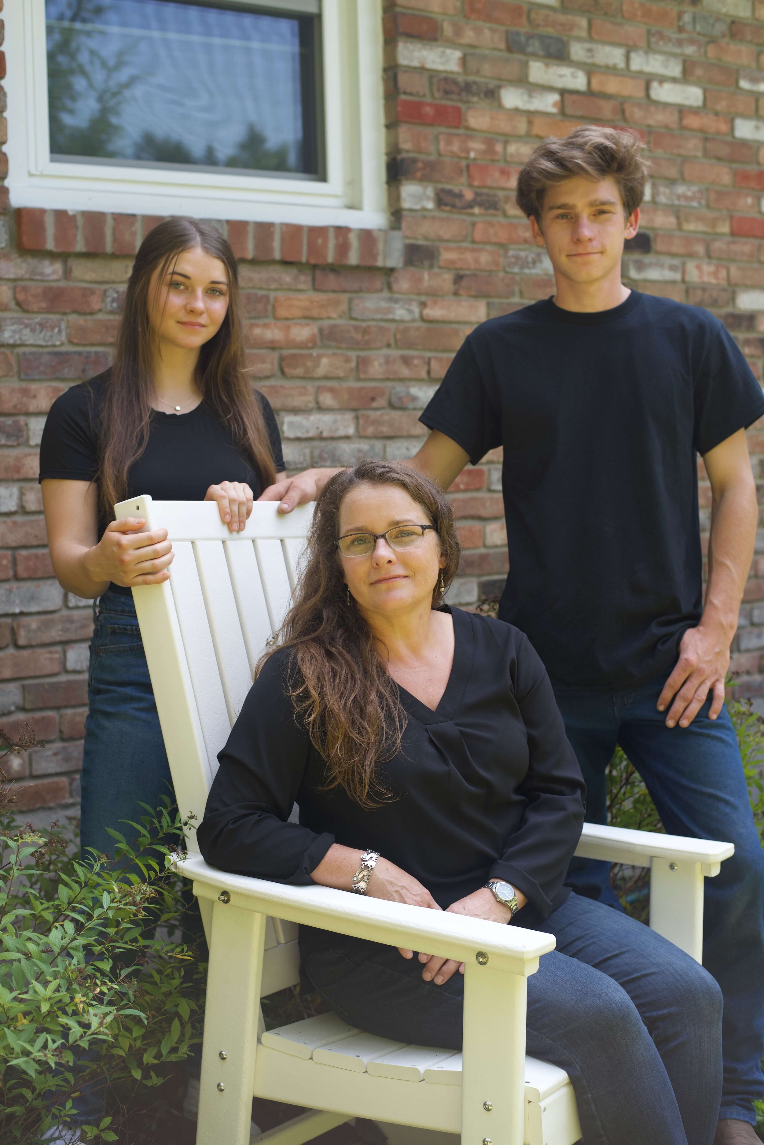 A woman with long brown hair sitting on a white chair outdoors, with two young people standing behind her, one male and one female, in front of a brick house. All three are dressed in black.