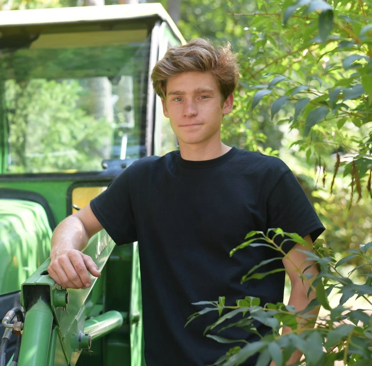 Young man wearing a black t-shirt standing outdoors near a green tractor surrounded by trees and foliage.