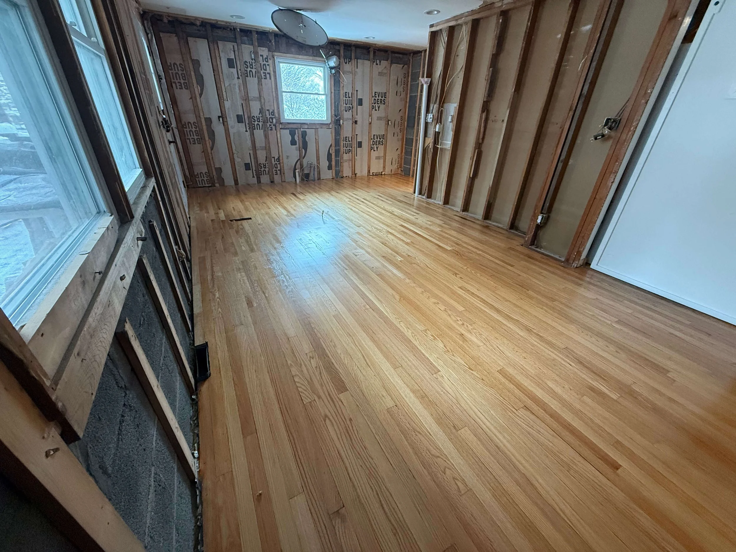 Empty room with hardwood floors, unfinished walls with exposed framing, a window and some electrical wiring are visible.