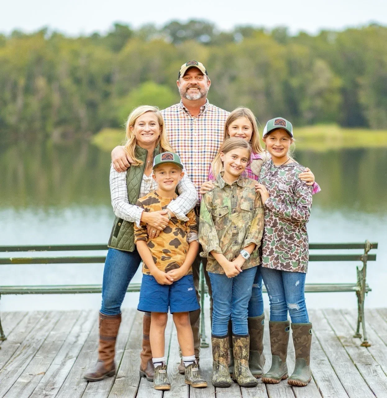 Family of six standing on a wooden dock near a lake with trees in the background, smiling for a photo. They are dressed in casual outdoor clothing, with some wearing camo and rain boots.