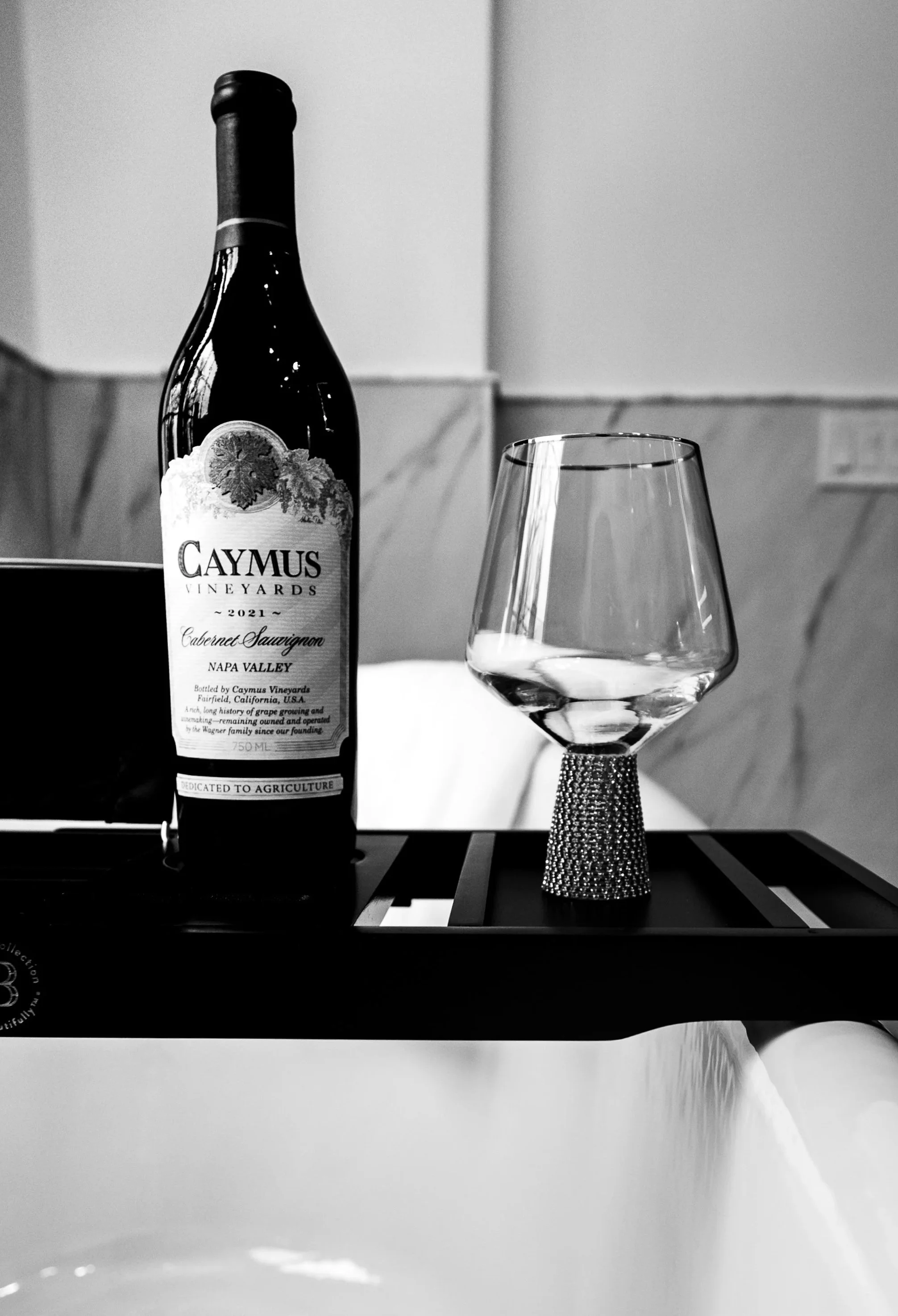 A black-and-white photo of a wine bottle labeled 'Caymus Vineyards 2021 Cabernet Sauvignon Napa Valley' and an empty wine glass with a textured base on a tray, on a countertop with a tiled wall in the background.