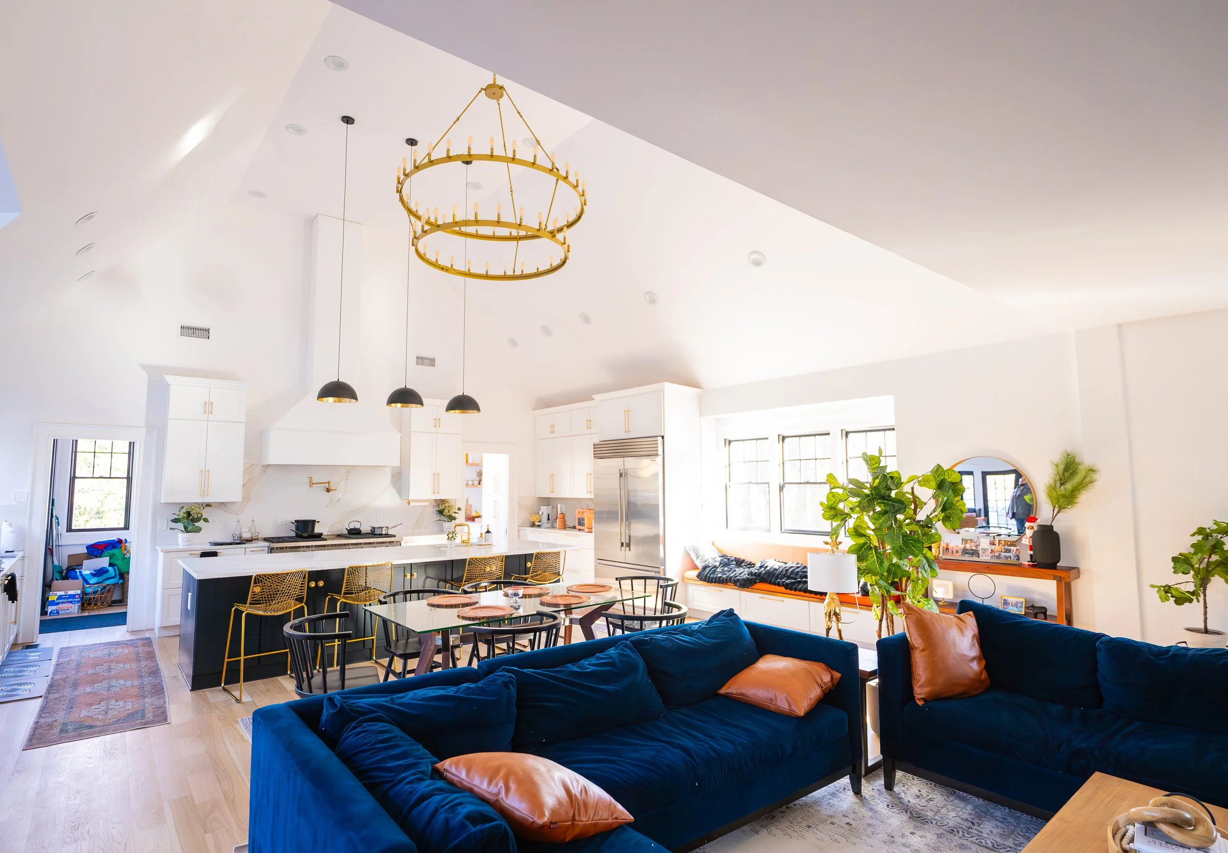 Bright open-concept living room and kitchen with white walls, high vaulted ceiling, blue sofas with brown pillows, green plants, and large windows.
