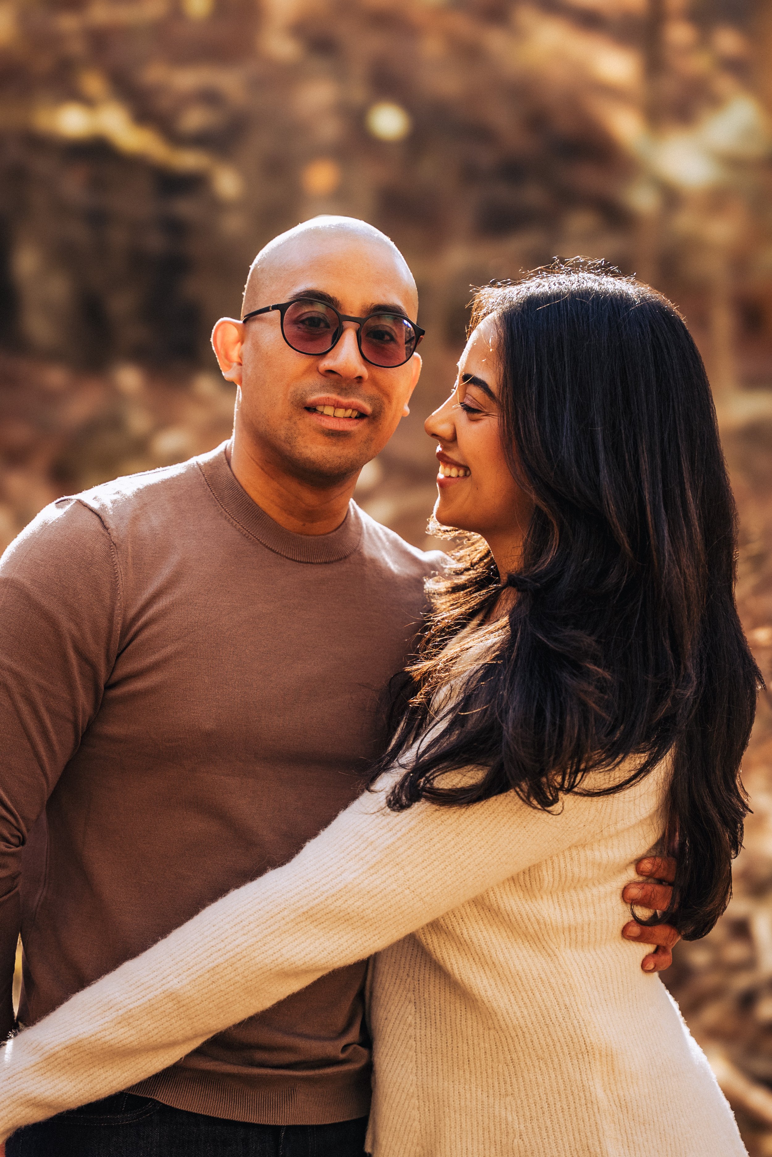 A man with bald head, wearing glasses and a brown shirt, hugging a woman with long dark hair, smiling, against a rocky outdoor background.