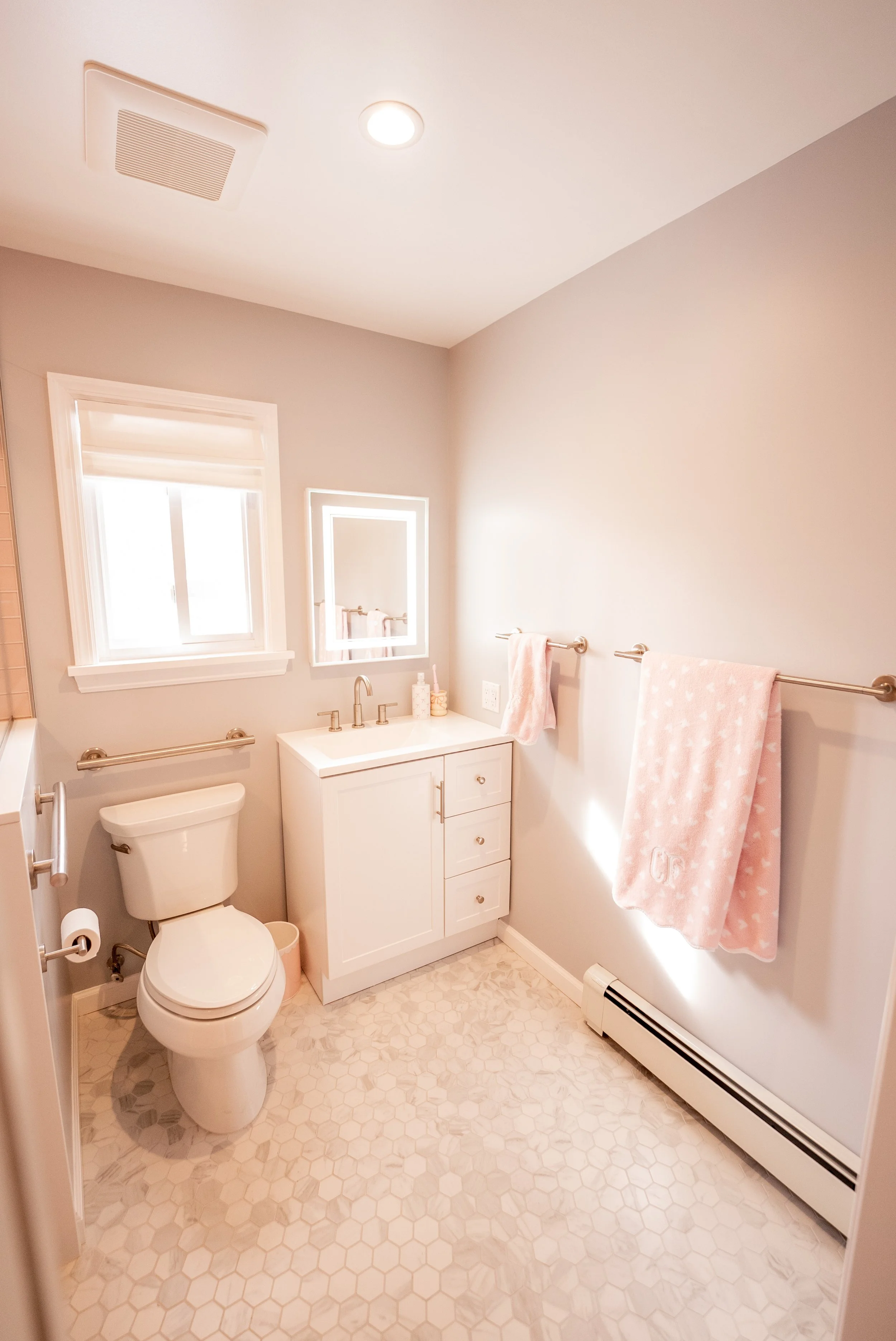 A small bathroom with a white toilet, a white vanity with a sink, a mirror with backlight, a window, and pink towels on bronze towel bars.