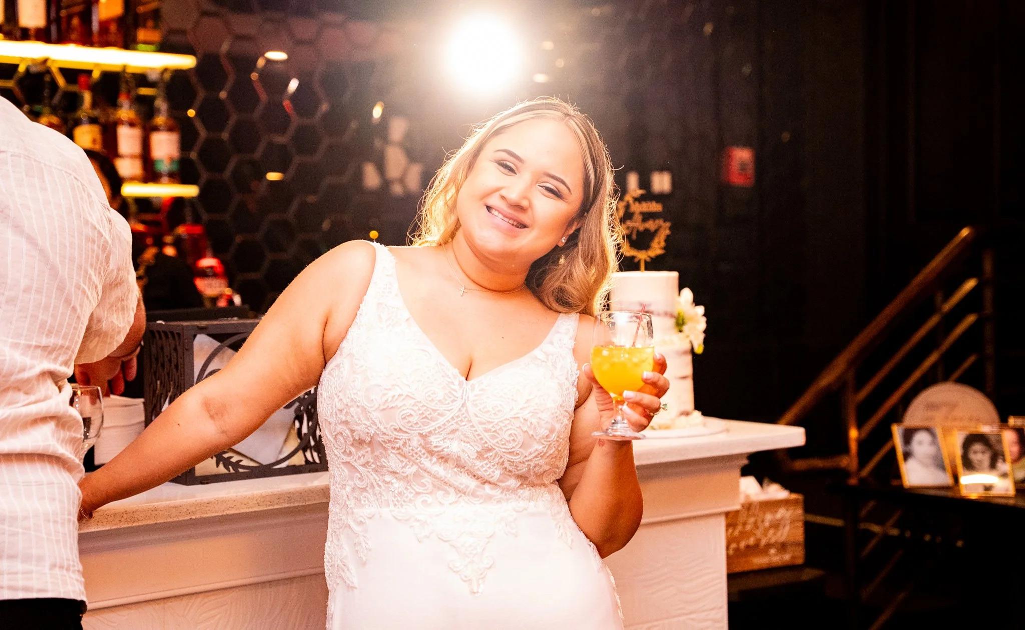 A woman in a white lace dress at a celebration, holding a yellow cocktail, smiling, with a birthday cake in the background, in a dimly lit venue.