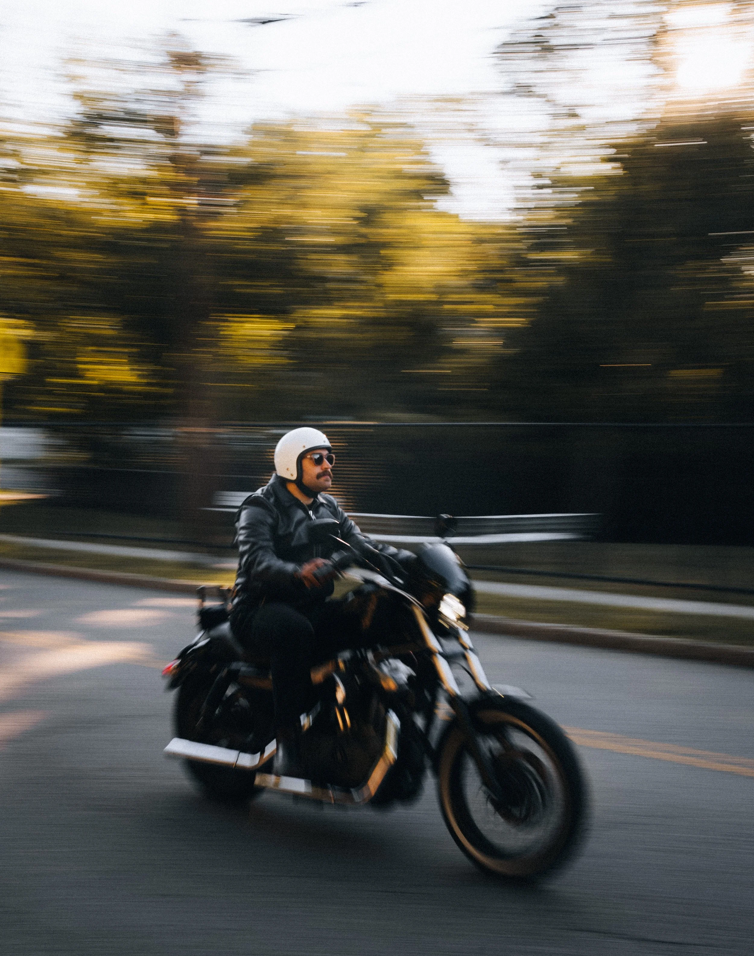 Man riding a black motorcycle on the road with trees and sunset in the background, wearing a white helmet and sunglasses.
