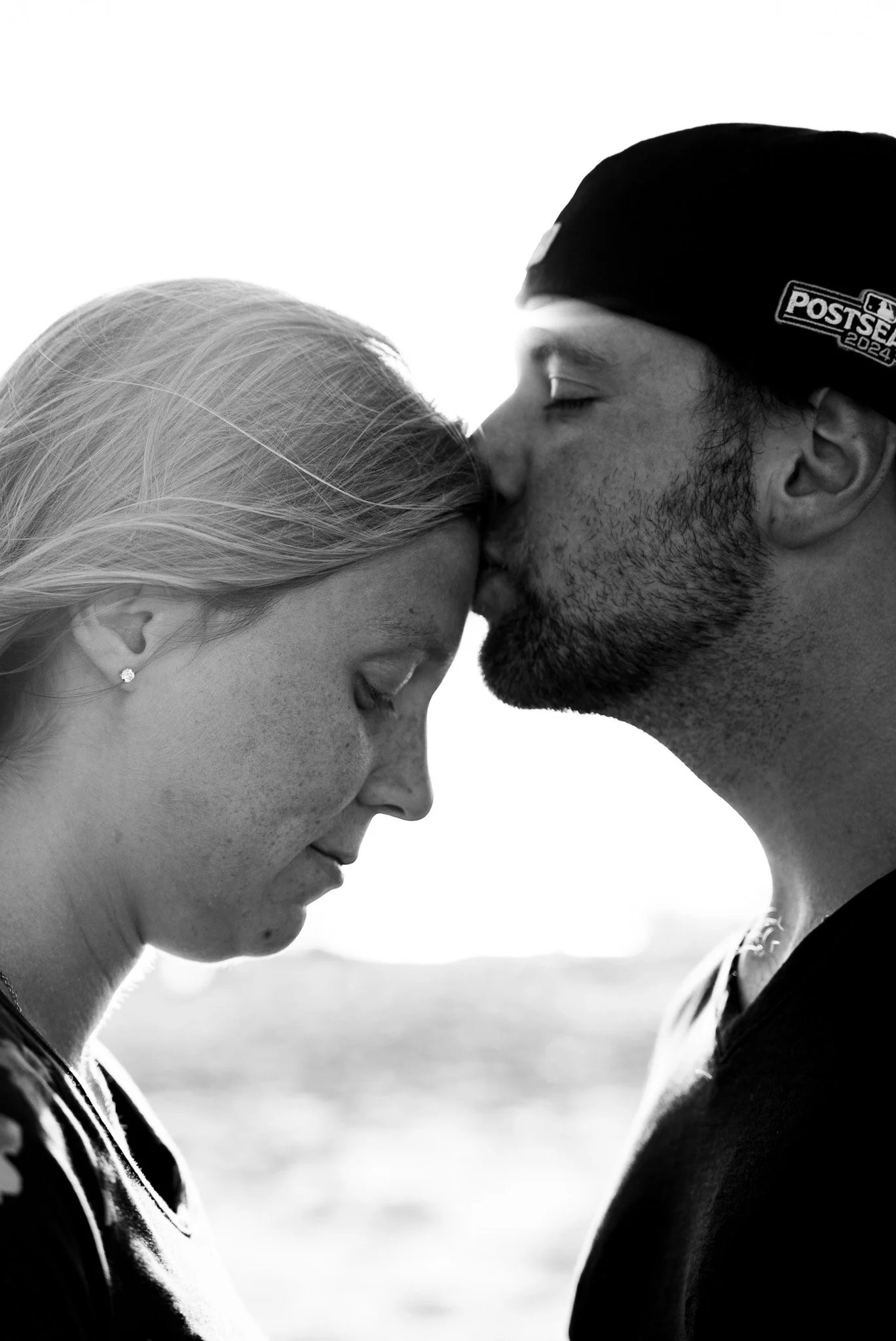 A black-and-white photograph of a couple with their foreheads touching and eyes closed; the woman has light hair and is wearing earrings, while the man has a beard, a cap with a logo, and freckles.