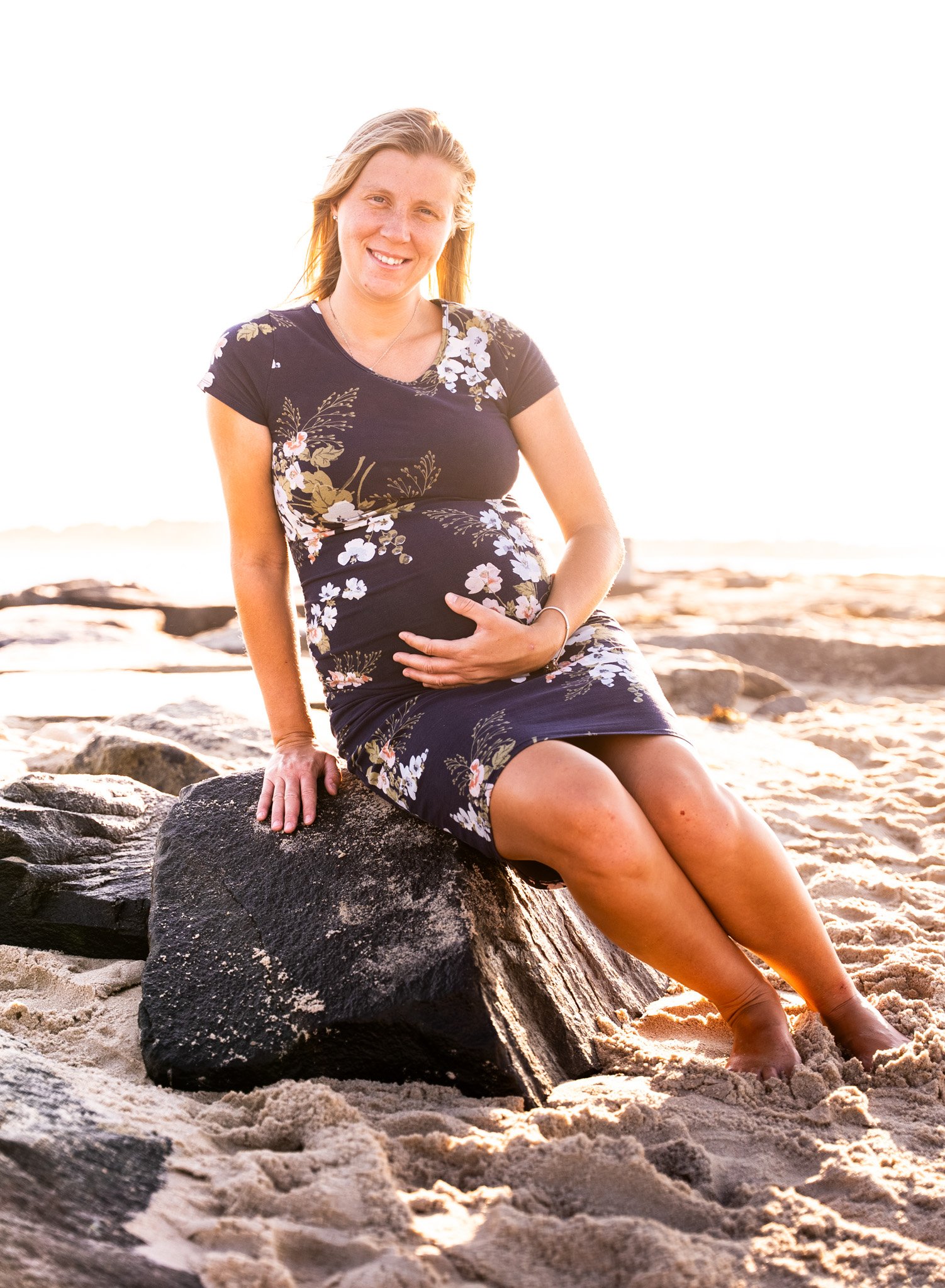 A pregnant woman sitting on a rock at the beach during sunset, smiling and holding her belly with one hand.