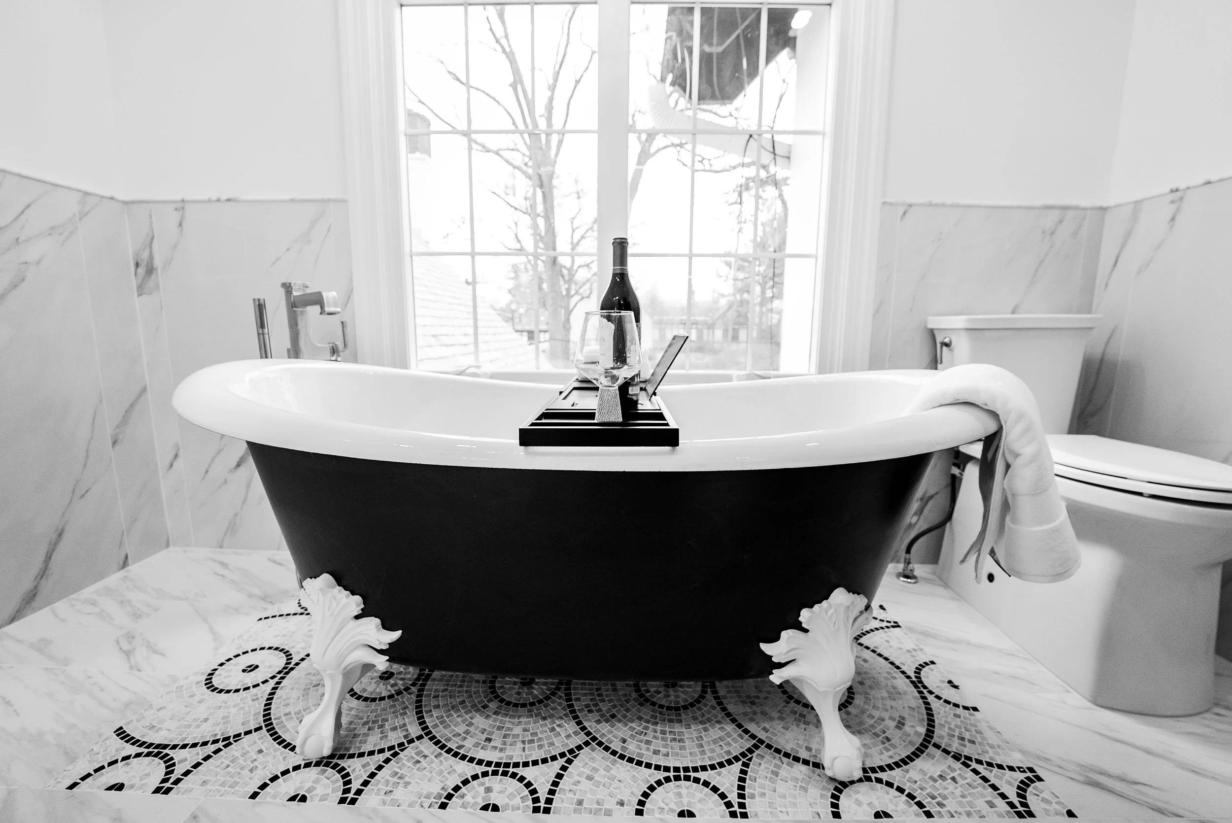 A black and white bathroom featuring a vintage clawfoot bathtub with a wine bottle, glass, and laptop on a tray, a large window with view of trees outside, and a toilet nearby.
