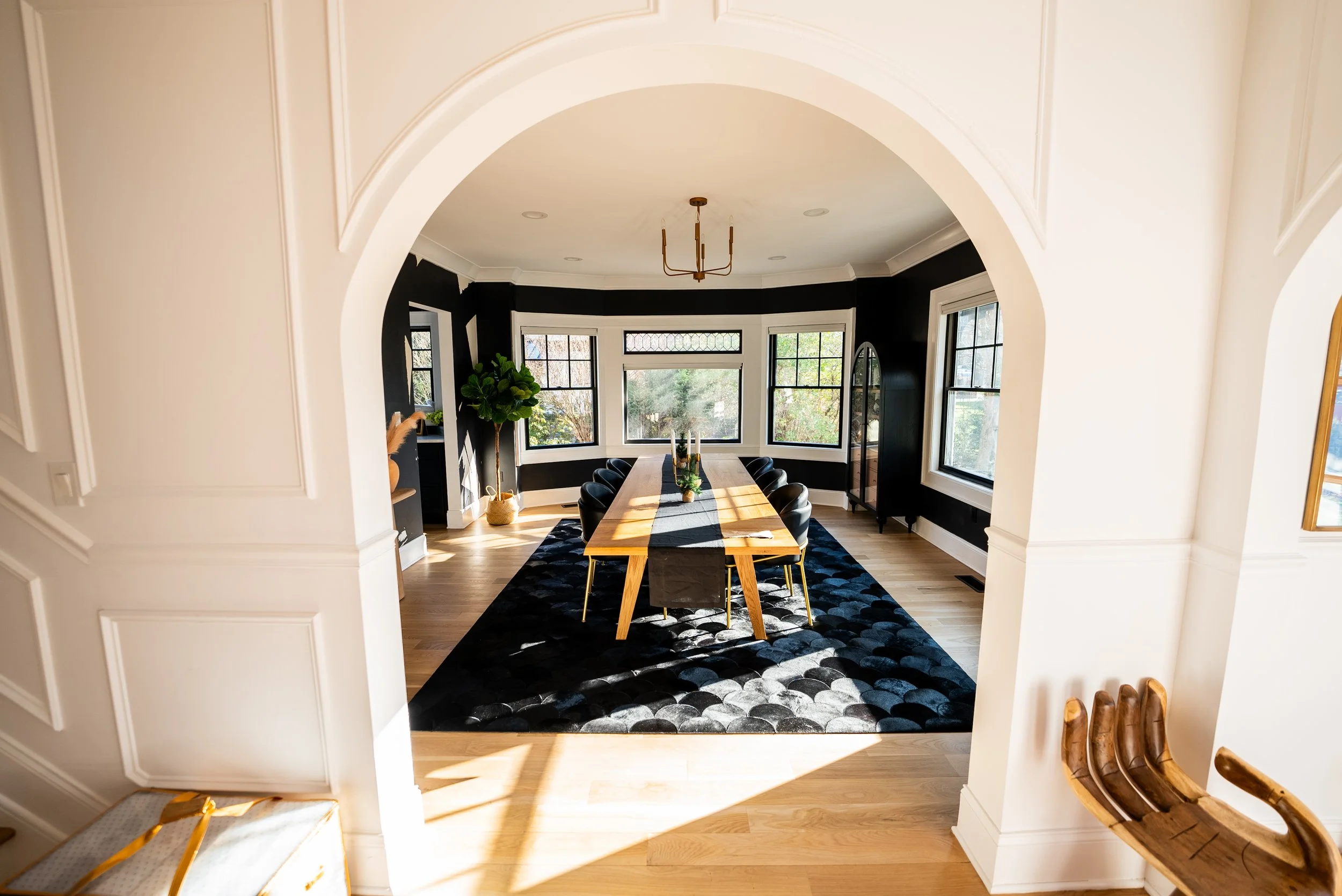 A modern dining room with a wooden table, black chairs, a black and gray patterned rug, and multiple windows letting in natural light. There's a tall green plant and a black display cabinet near the windows.