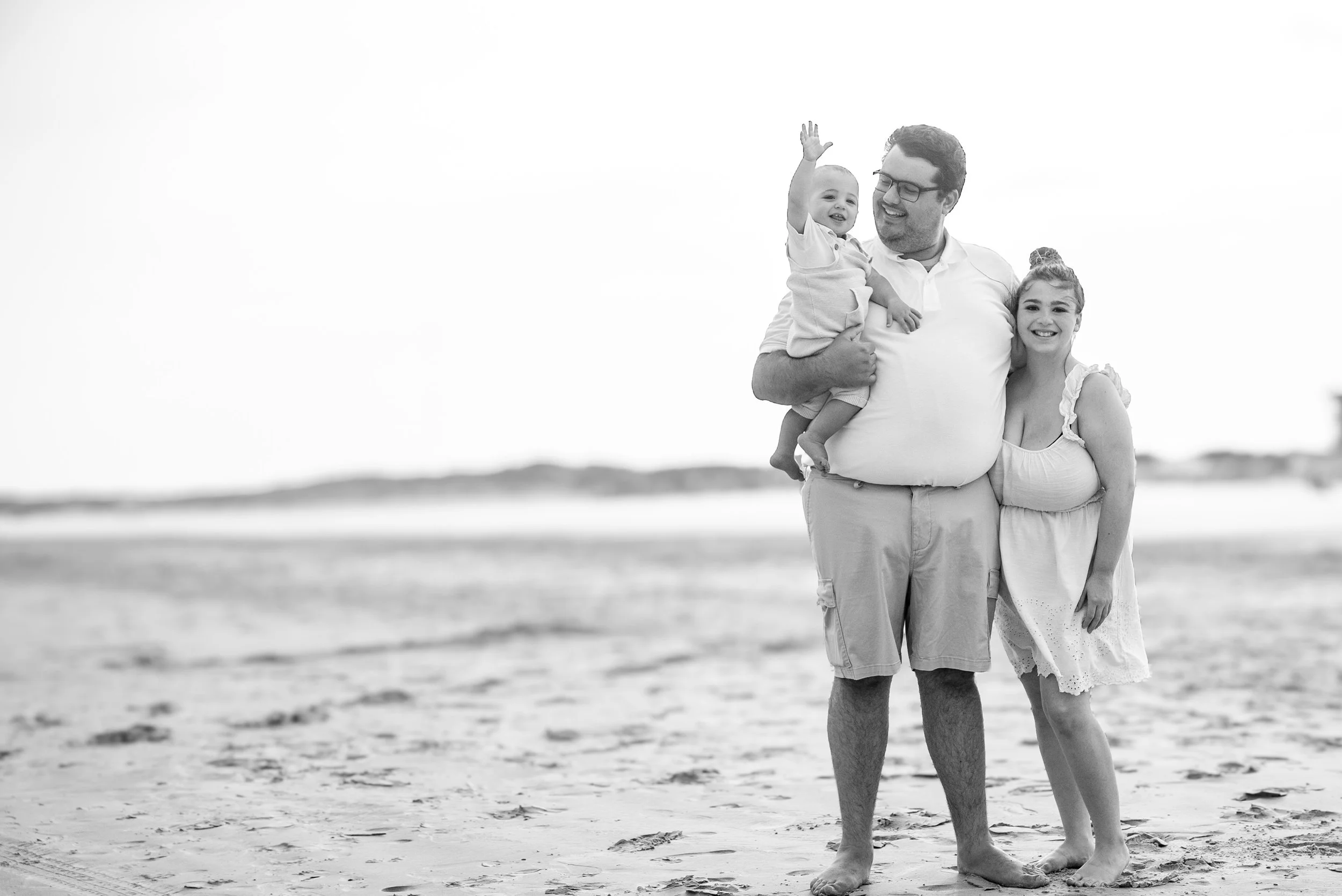 A family of three standing on a beach, smiling and enjoying their time together, with the father holding a young child and the mother standing beside them.