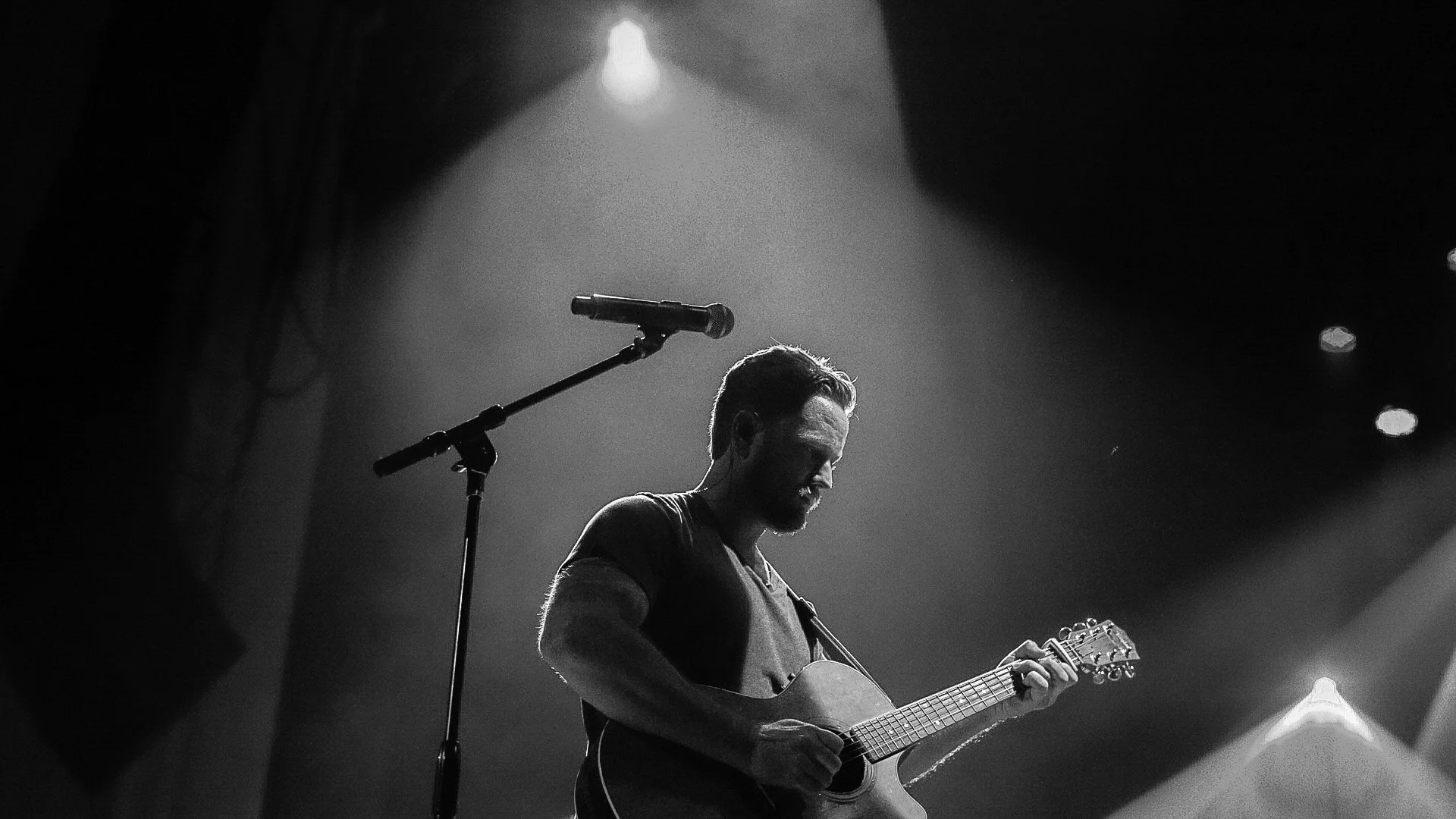 A musician playing an acoustic guitar on stage under a spotlight, with a microphone in front, in black and white.
