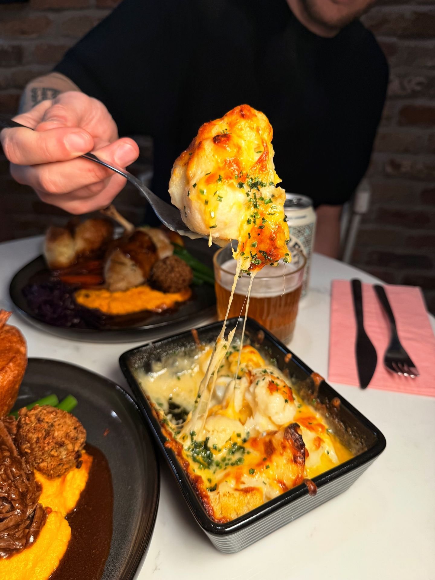 Person holding a spoon with cheesy baked casserole or potato dish over a table of various foods, including a plate of meatballs with vegetables, and a glass of beer.