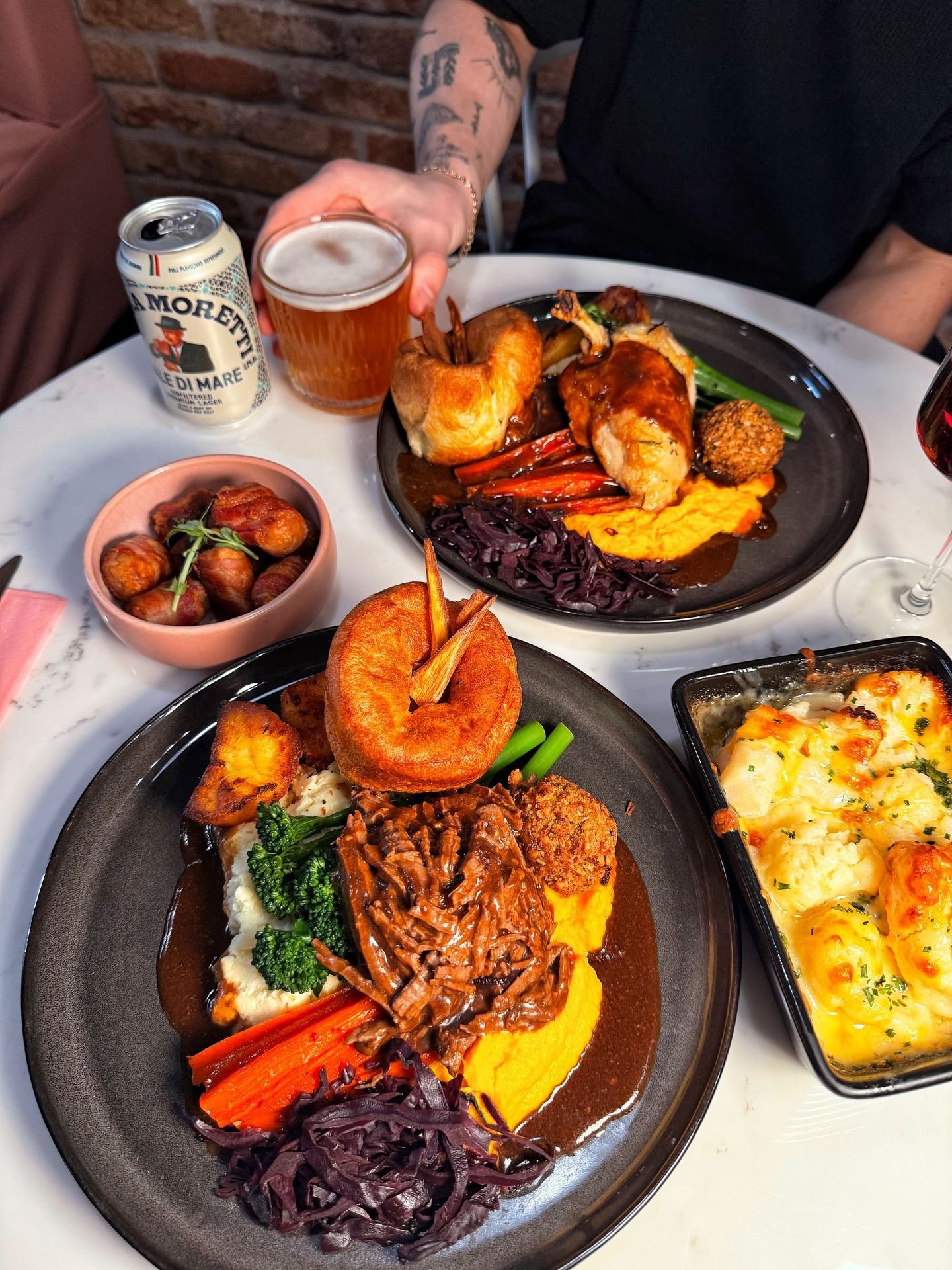 Two plates of hearty beef stew with mashed potatoes, vegetables, and roasted potatoes, with a side dish of cheesy baked casserole, and drinks including beer and wine on a white table.