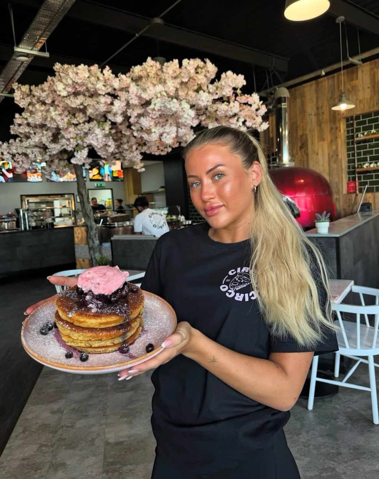 A woman with blonde hair holding a plate of stacked pancakes topped with pink ice cream and berries, inside a restaurant with a flower tree in the background.