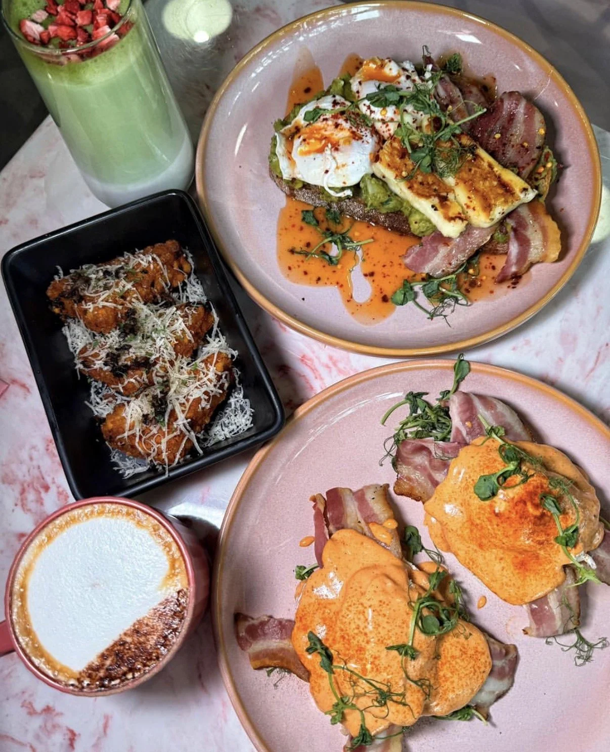 A meal with two plates of eggs Benedict topped with sauce and herbs, a black rectangular dish of fried chicken pieces or gnocchi with cheese, a glass of green smoothie or juice, and a cup of creamy coffee or dessert, all on a pink and white marble ta