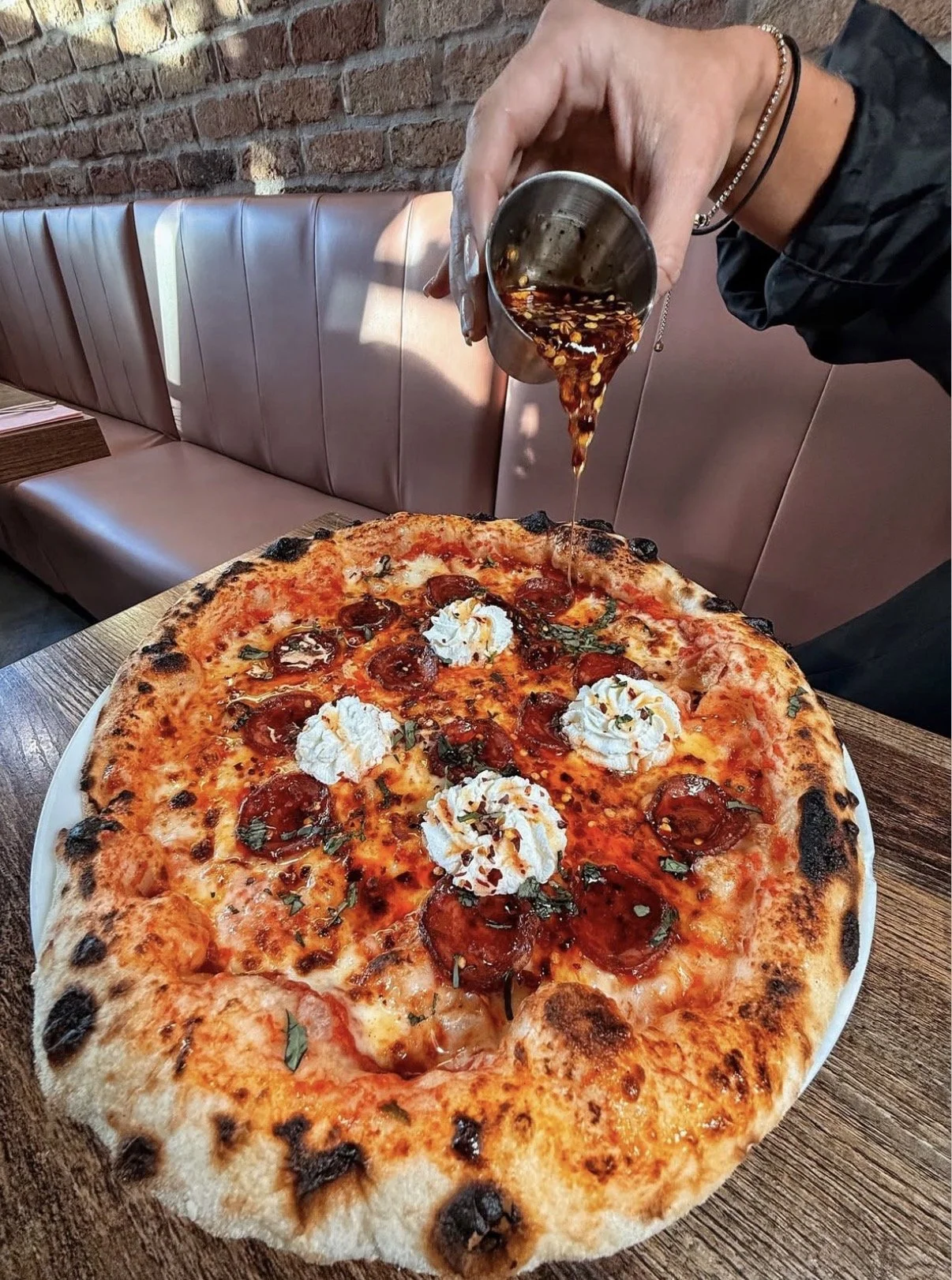 Person pouring balsamic glaze over a pepperoni pizza with dollops of ricotta cheese on a wooden table in a restaurant with brick wall background.