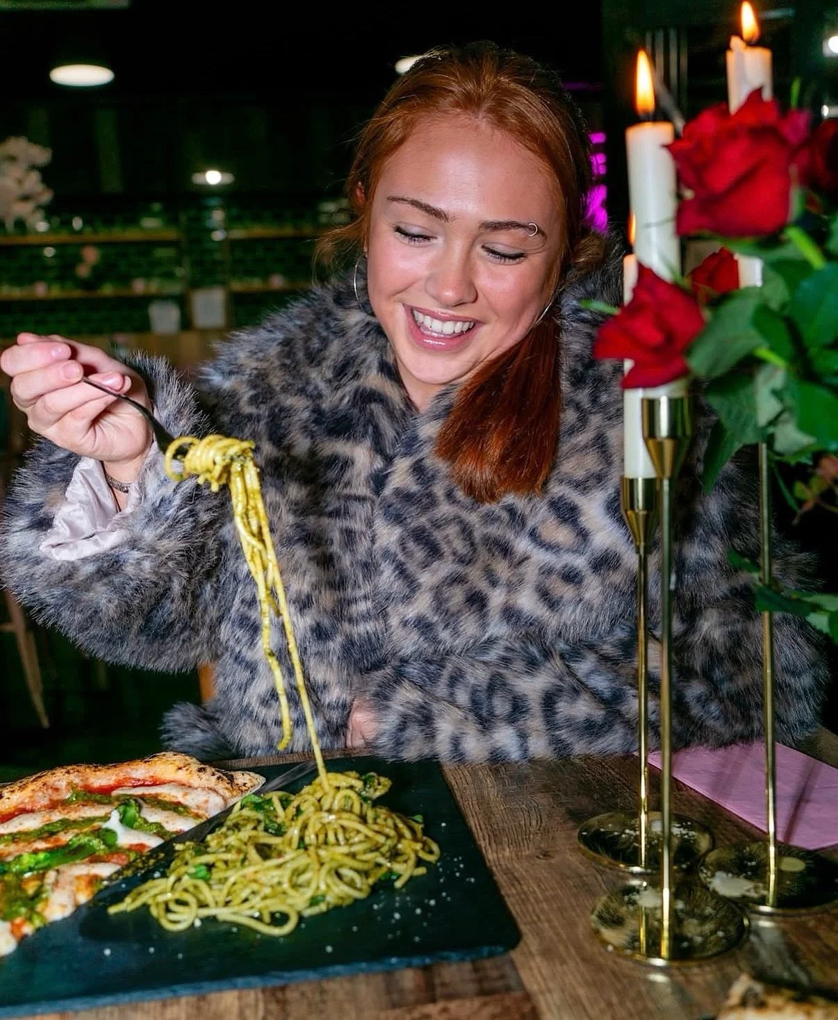A smiling woman wearing a leopard print fur coat is enjoying spaghetti pasta at a restaurant table with candles and red roses.