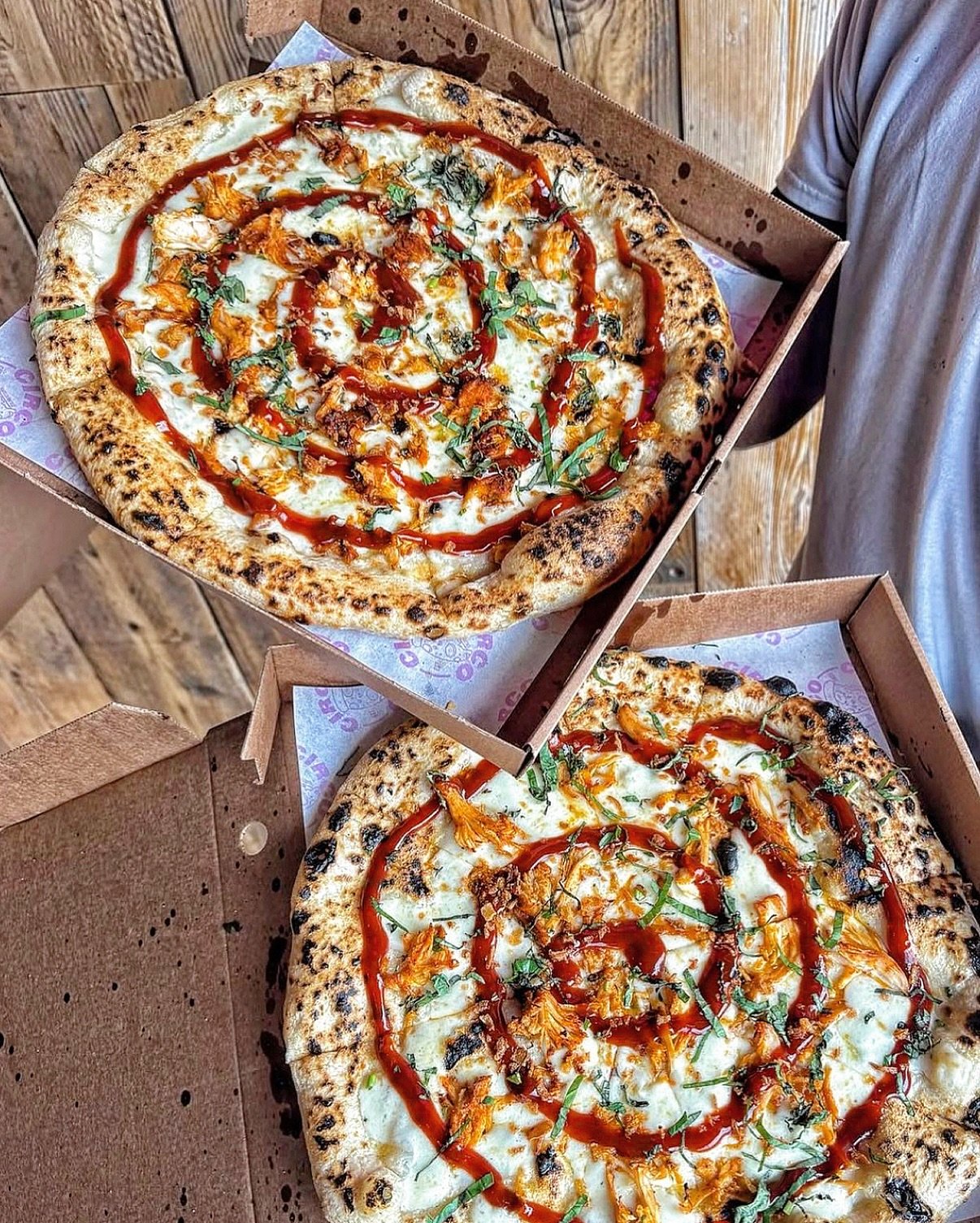 Two pizzas with cheese, toppings, and a spiral pattern of hot sauce inside a cardboard pizza box.