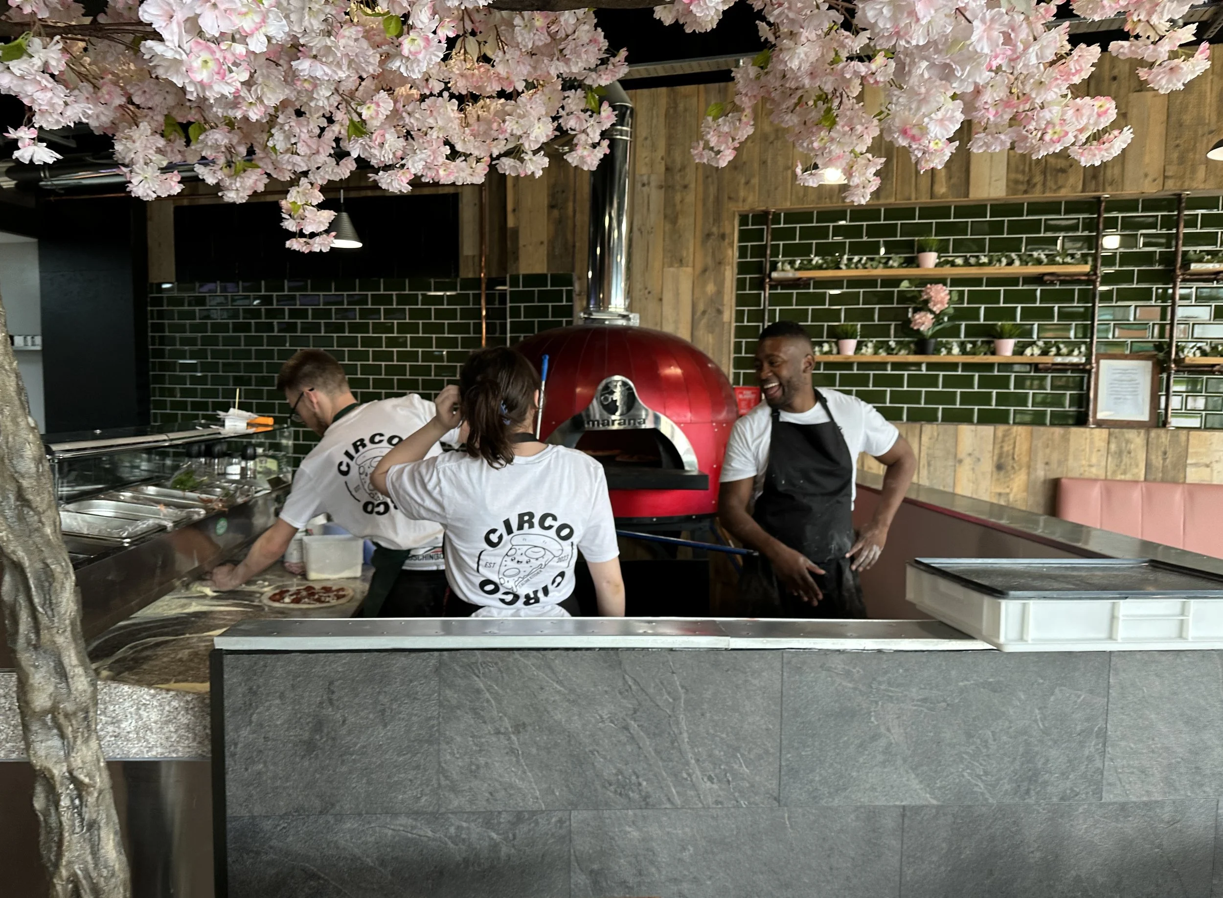 Three staff members working at a pizza restaurant, with a red pizza oven, green tiled wall, and pink flowers hanging from above.