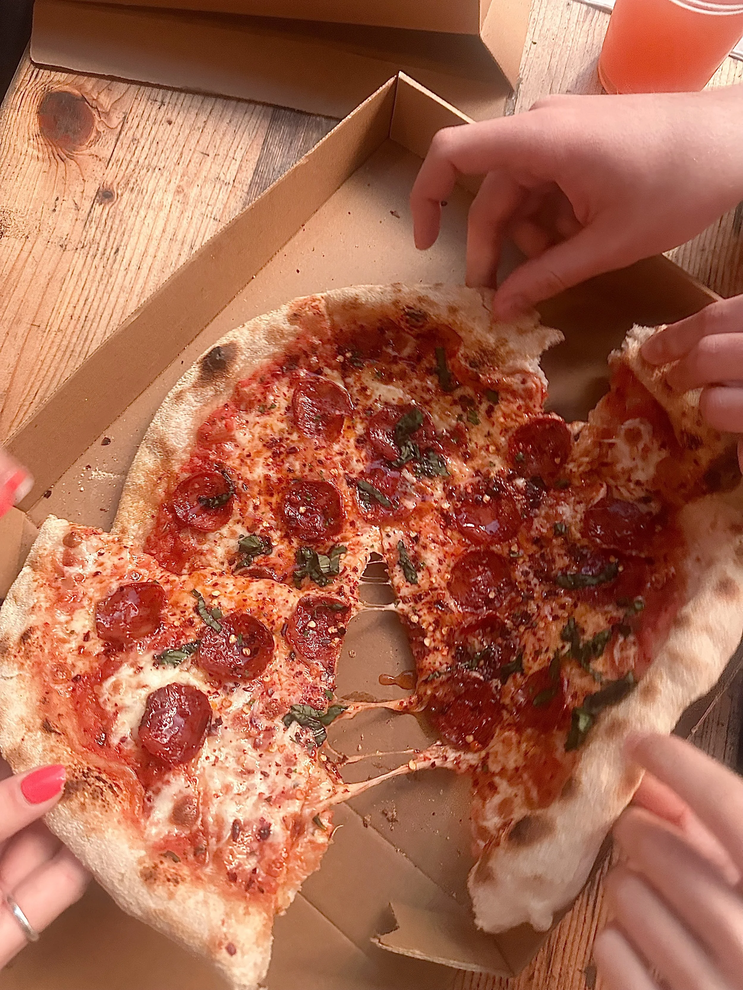 People grabbing slices from a pepperoni and basil pizza in a cardboard box